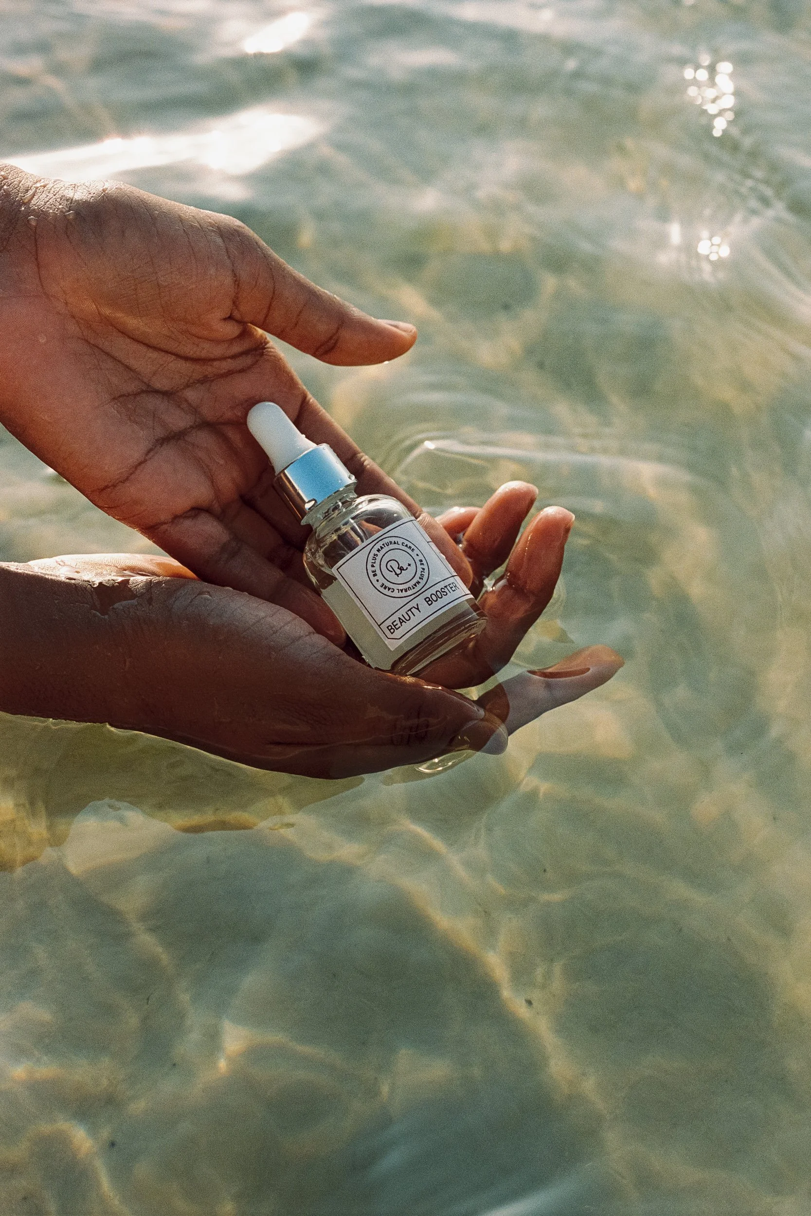 Wet hands cradling a Beauty Booster dropper bottle in clear lagoon water, underwater, Be Plus Natural Care, Lençóis Maranhenses, Brazil, on 35mm film.