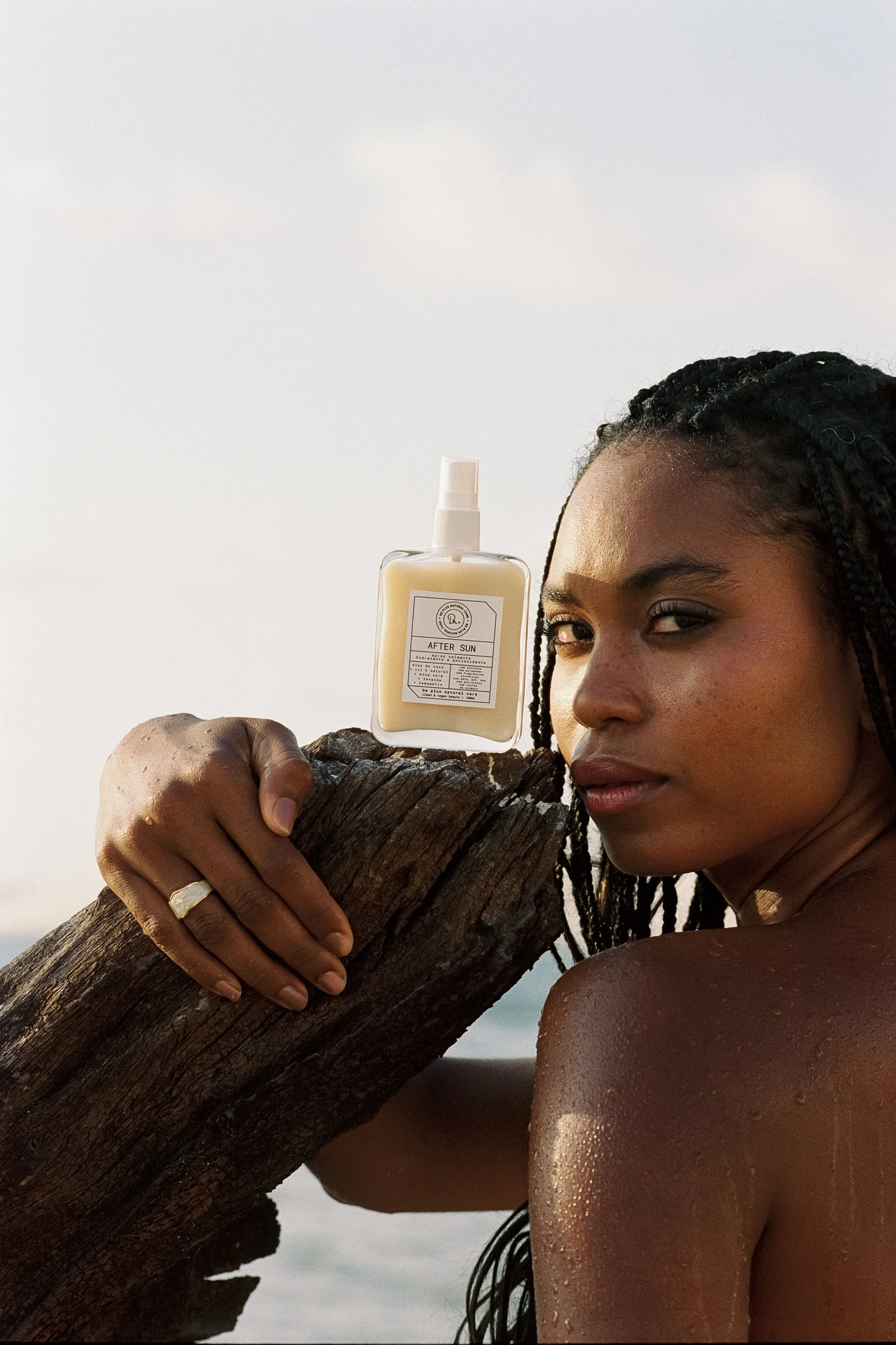 Model looking over her wet shoulder beside After Sun spray set on driftwood, Be Plus Natural Care, wet skin, sunset, Lençóis Maranhenses, Brazil, on 35mm film.