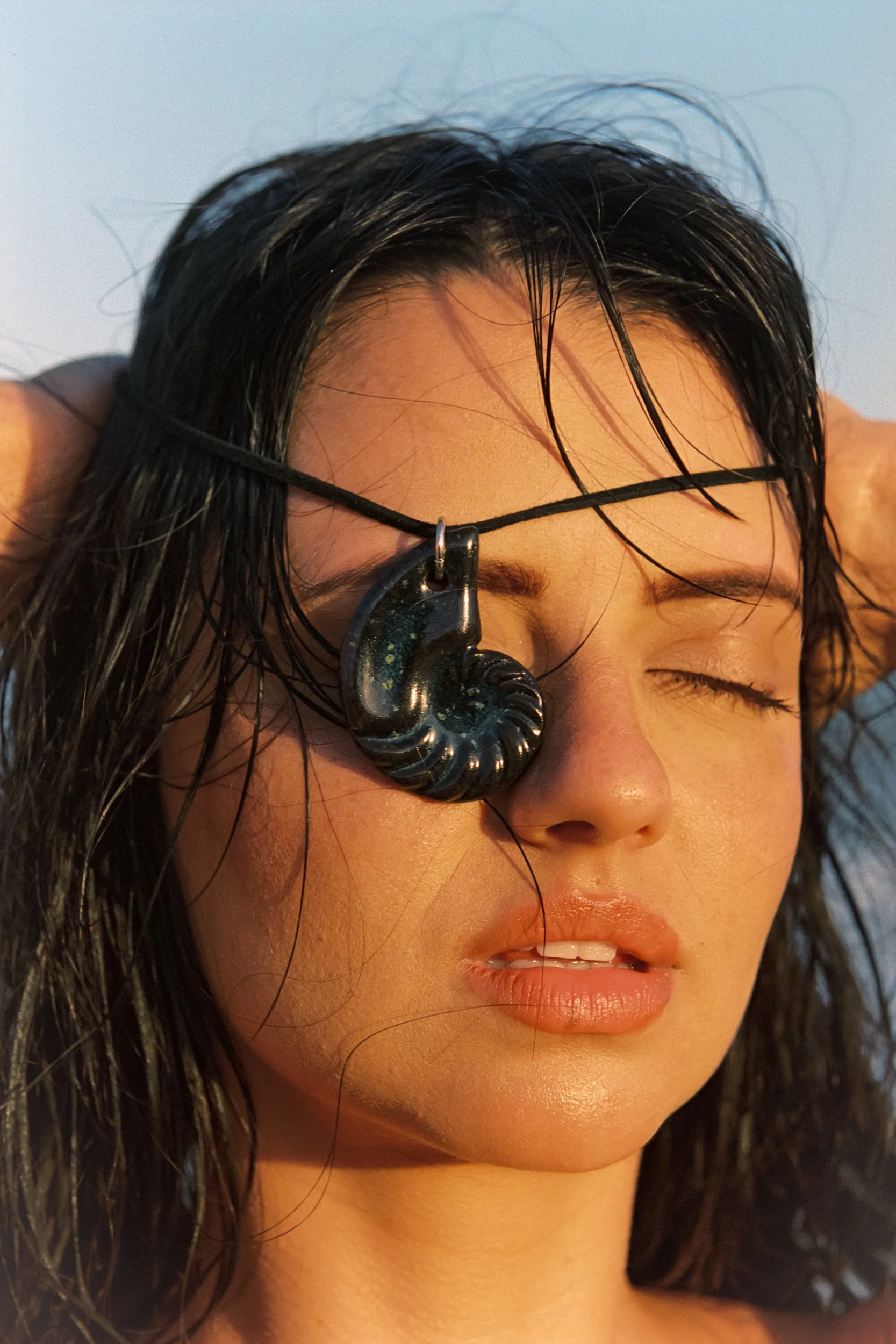 Tight crop of shell pendant over eye, lips parted, blue sky, 35mm film