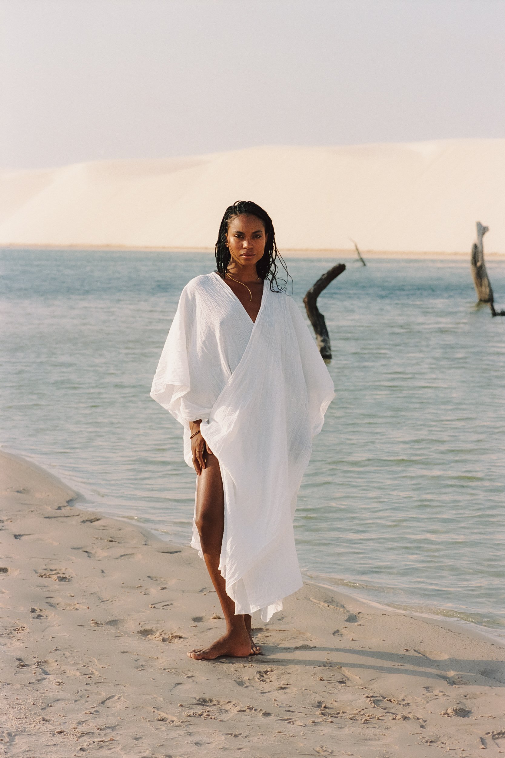Full-length view of a model barefoot on lagoon sandbar in a white kaftan, Be Plus Natural Care, Lençóis Maranhenses, Brazil, on 35mm film.