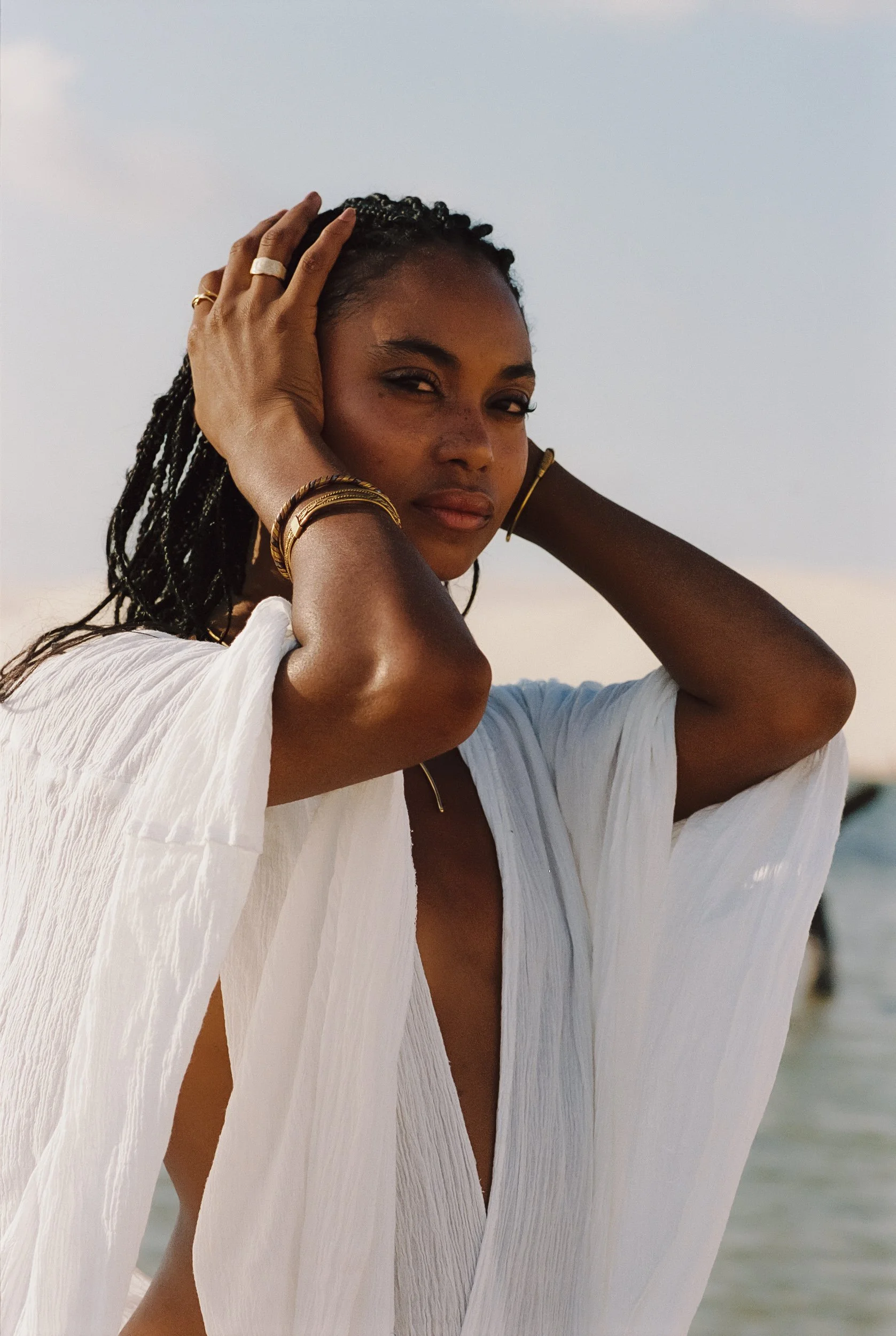 Model with both hands in her braids, gold bangles and open white draped kaftan, lagoon behind, Be Plus Natural Care, Lençóis Maranhenses, Brazil, on 35mm film.