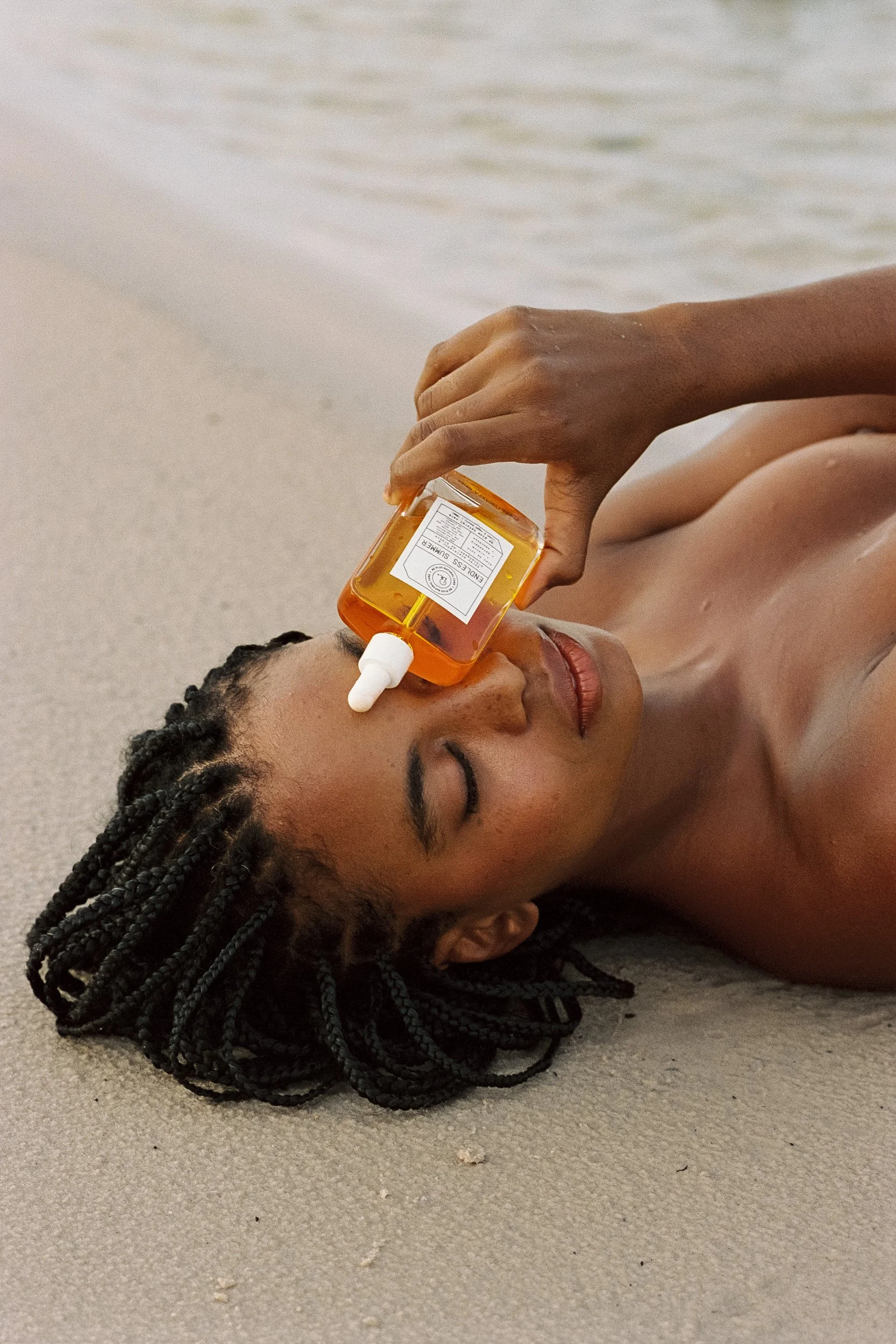 Model lying on the sand with eyes closed, resting an Endless Summer oil bottle on her face, Be Plus Natural Care, at golden hour, Lençóis Maranhenses, Brazil, on 35mm film.