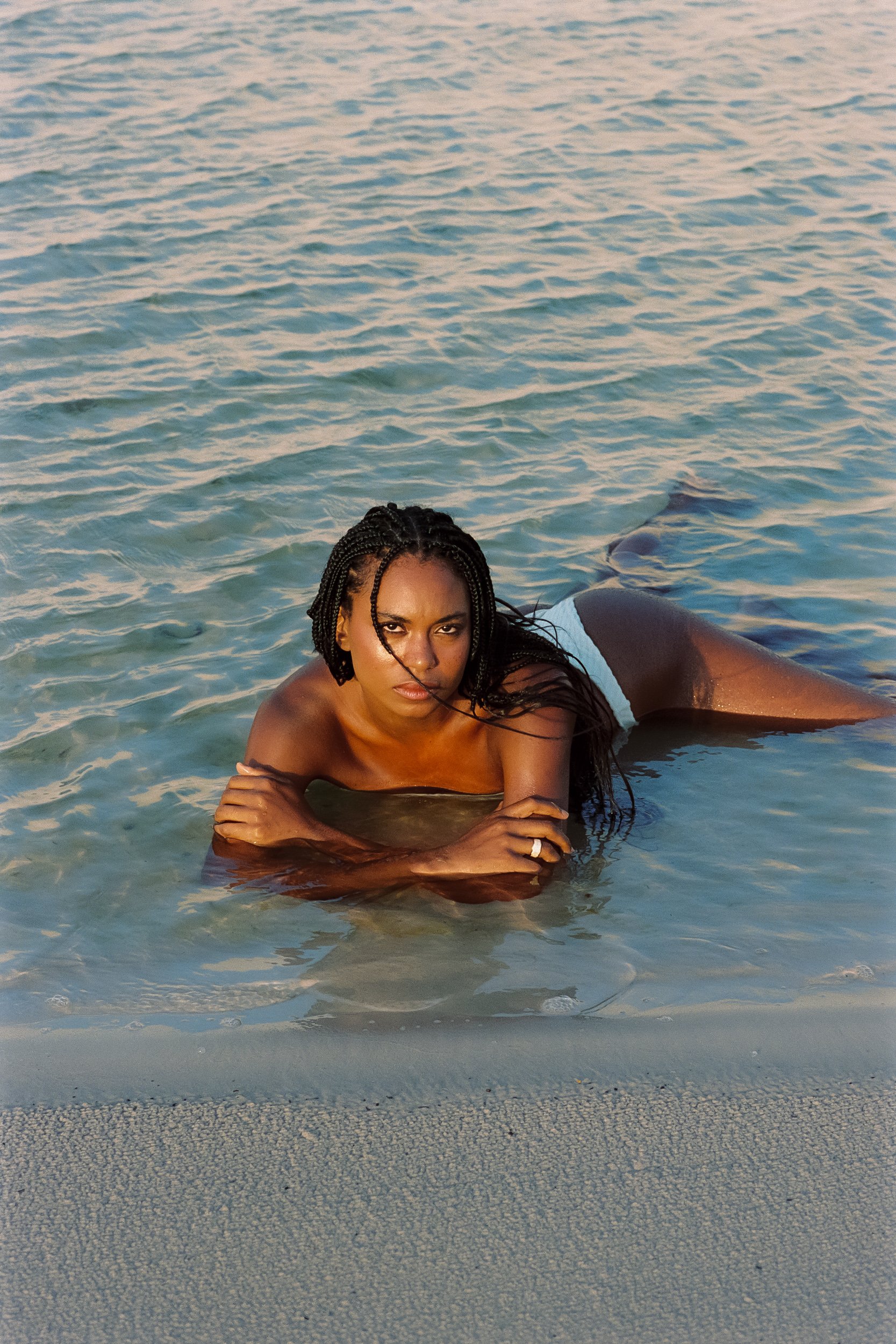 Model lying in shallow turquoise lagoon water, arms crossed on the sand at golden hour, Be Plus Natural Care, Lençóis Maranhenses, Brazil, on 35mm film.