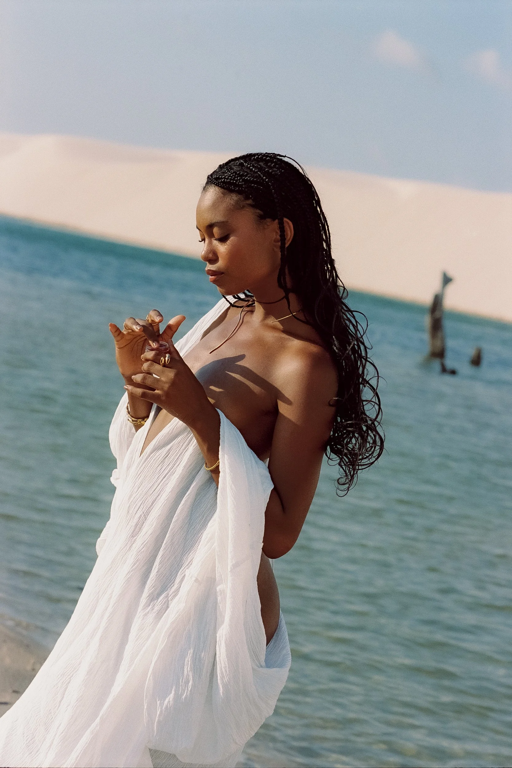 Model wrapped in white cotton dress holding a pink balm jar at her shoulder beside turquoise lagoon, Be Plus Natural Care, Lençóis Maranhenses, Brazil, on 35mm film.