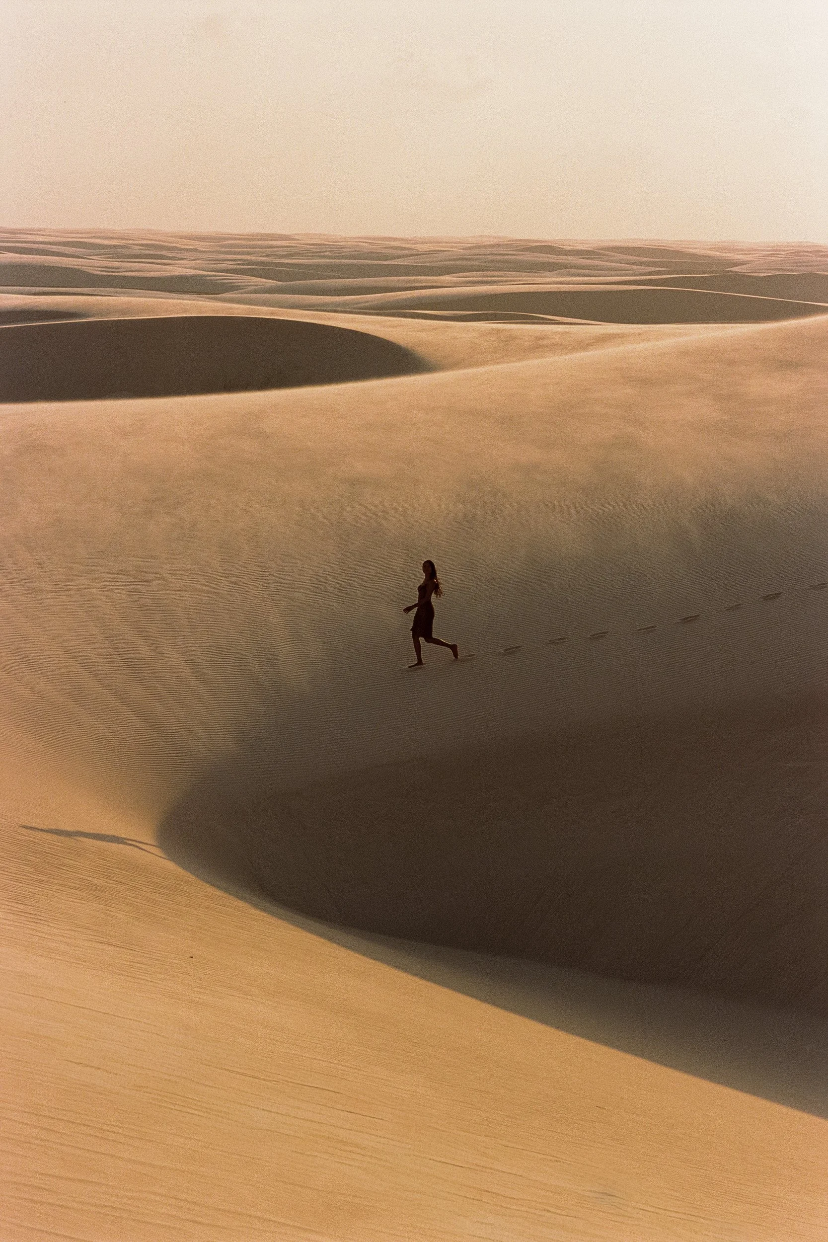 Small figure running barefoot across vast golden dunes, footprints trailing behind her, Ranaco Atelier, Lençóis Maranhenses, Brazil, on 35mm film.