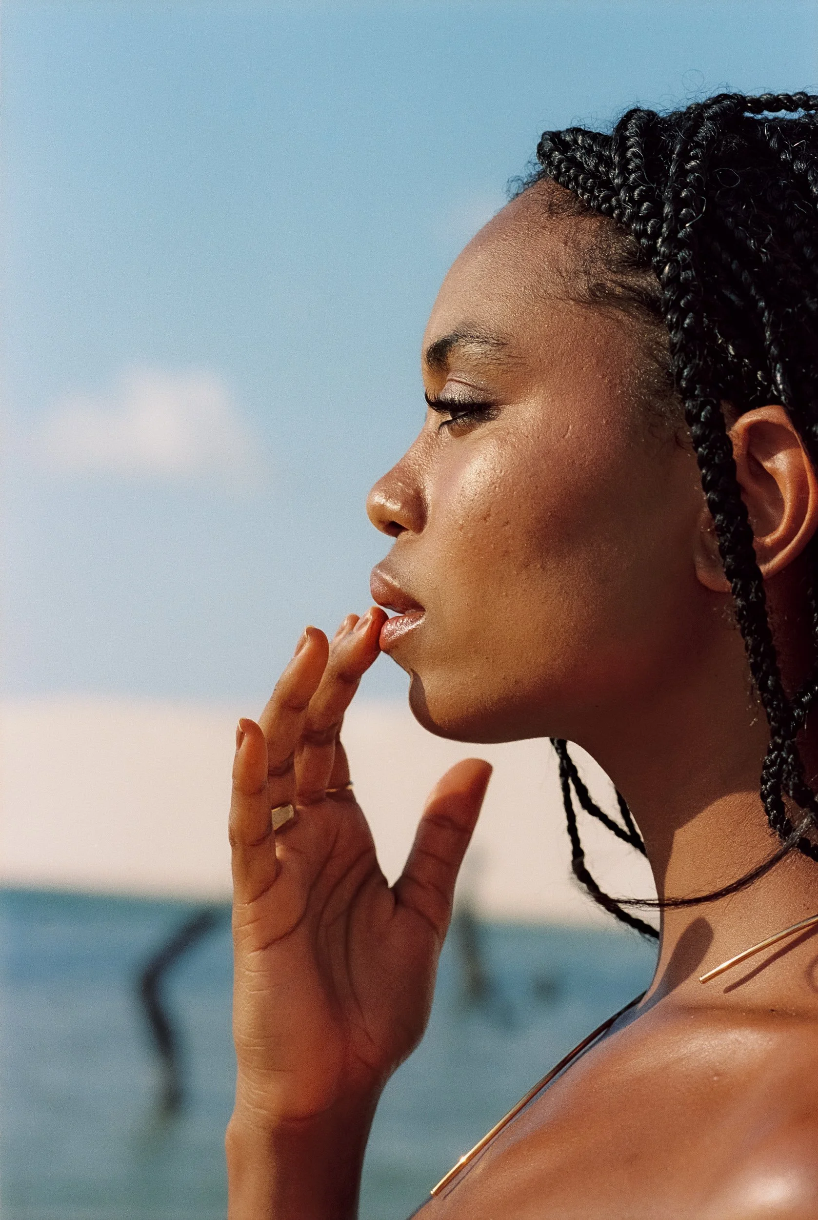 Close-up profile beauty portrait of a model touching her lips, braids pulled back, blue sky, dunes behind, Be Plus Natural Care, Lençóis Maranhenses, Brazil, on 35mm film.