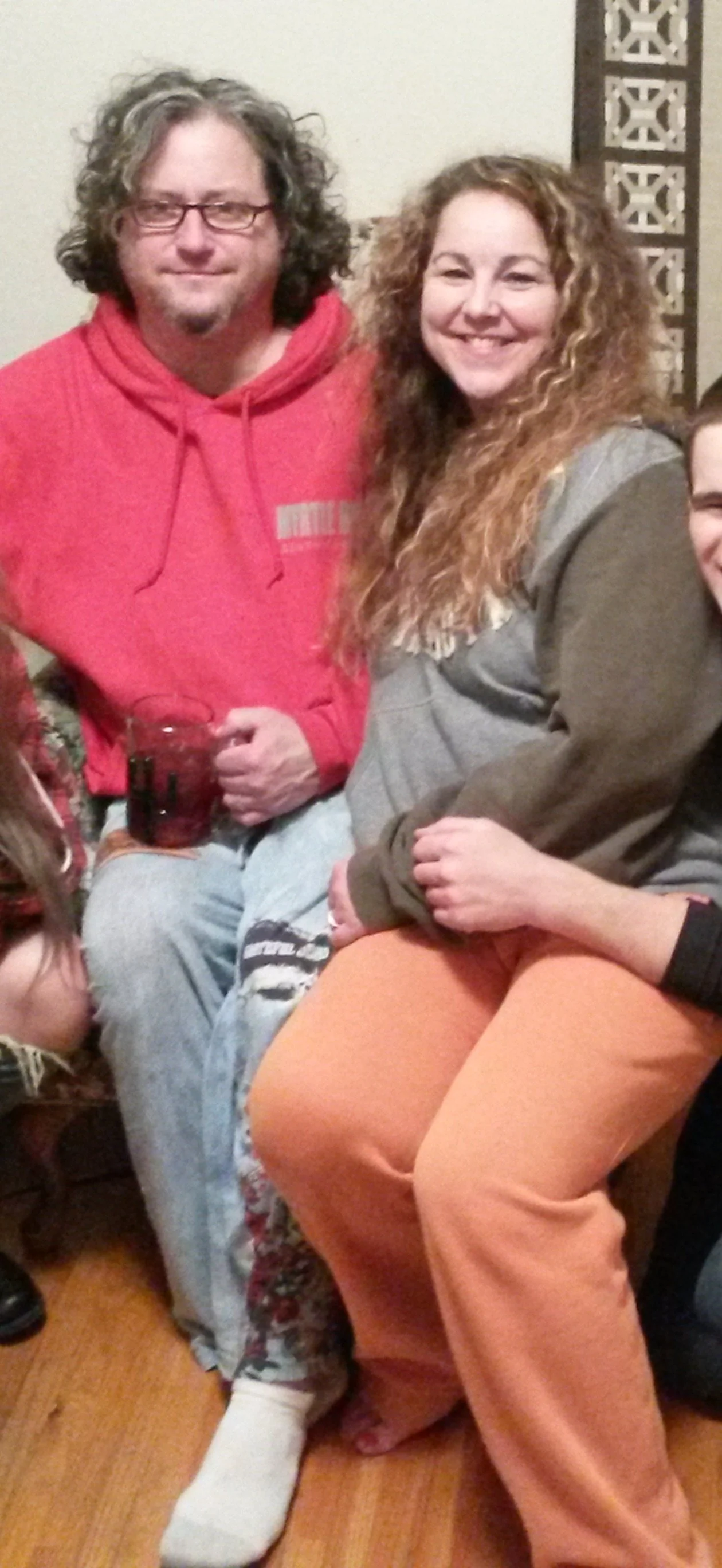 A man with curly hair and glasses wearing a red hoodie, and a woman with curly hair in a gray jacket and orange pants, smiling and sitting together indoors.