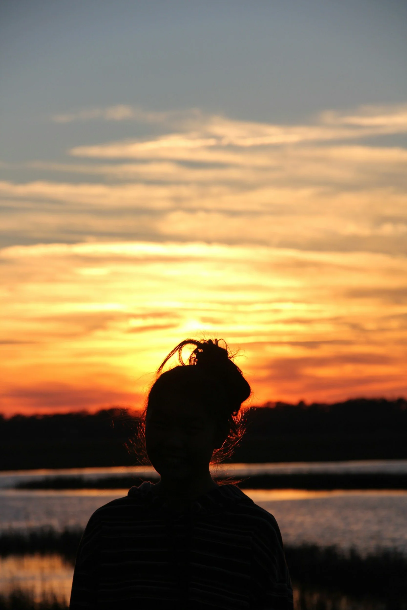 Silhouette of a person with hair tied up, sitting outdoors during sunset or sunrise, with clouds in the sky and a body of water in the background.