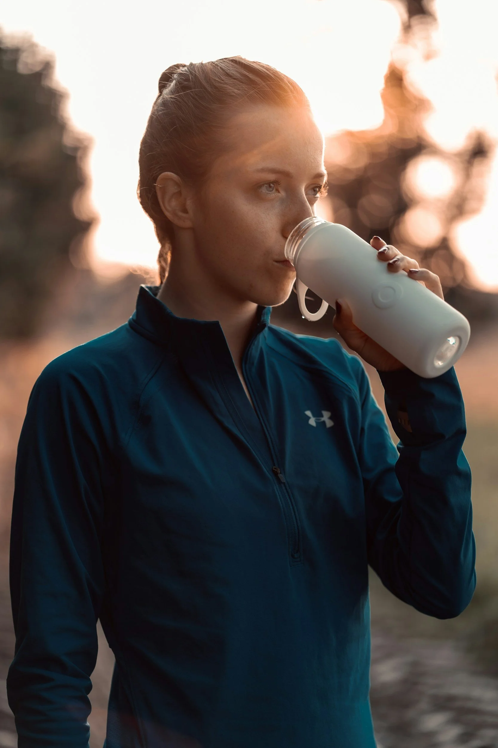 A young woman with red hair tied back, wearing a blue Under Armour athletic jacket, drinking from a white water bottle outdoors during sunset with trees in the background.