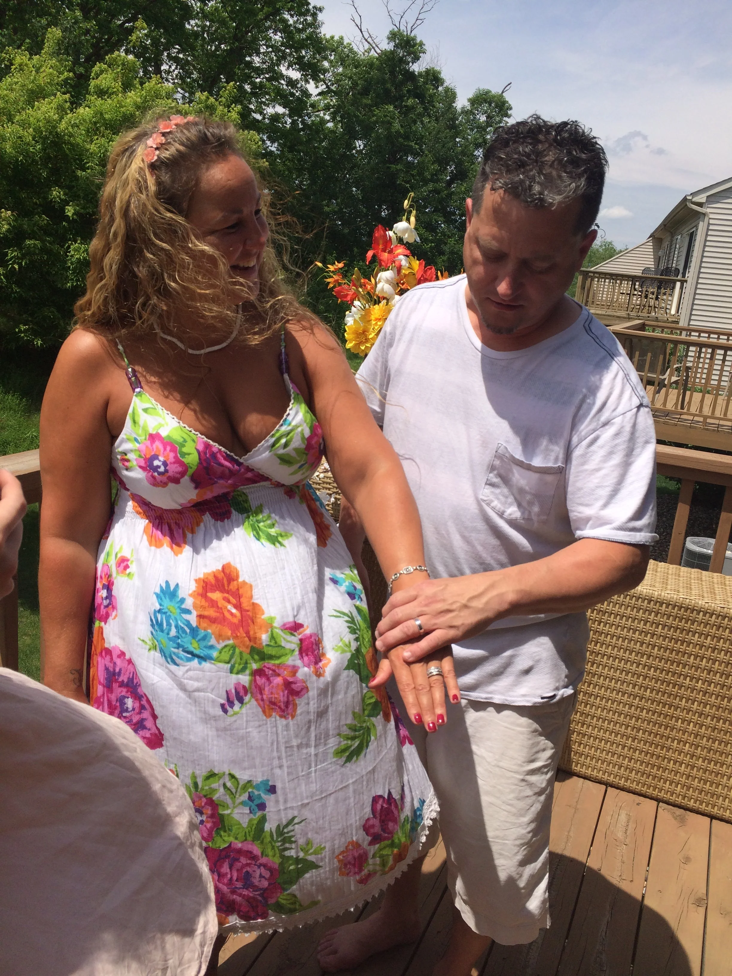 A woman in a colorful floral dress proudly shows off her pregnant belly while standing outside on a wooden deck with a man in casual white clothing. The woman is smiling and wearing a pink flower headband, and the man is looking at her belly with a serious expression. There are lush green trees and a house with a balcony in the background.