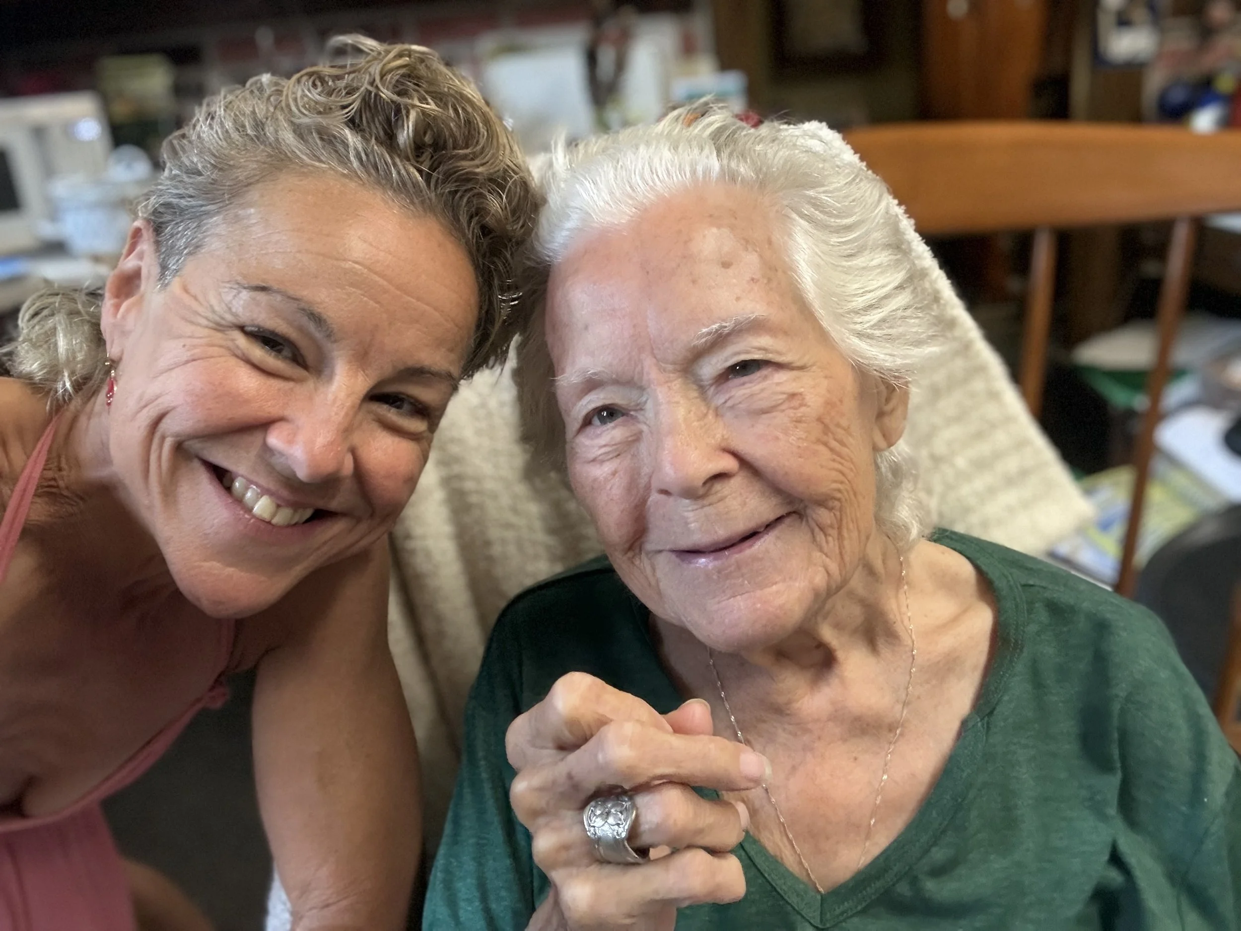 Two women, one elderly with white hair and one middle-aged with curly blonde hair, are smiling and posing closely together in a warm, domestic setting.