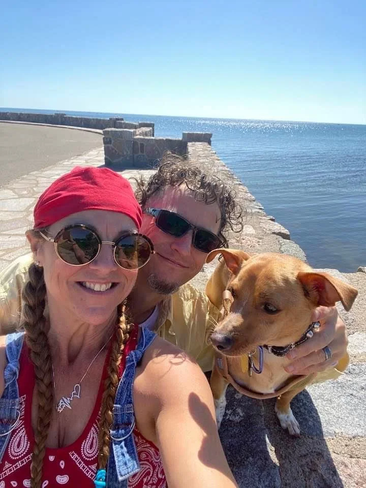 Two people smiling while taking a selfie near a stone pier by the water with their dog.