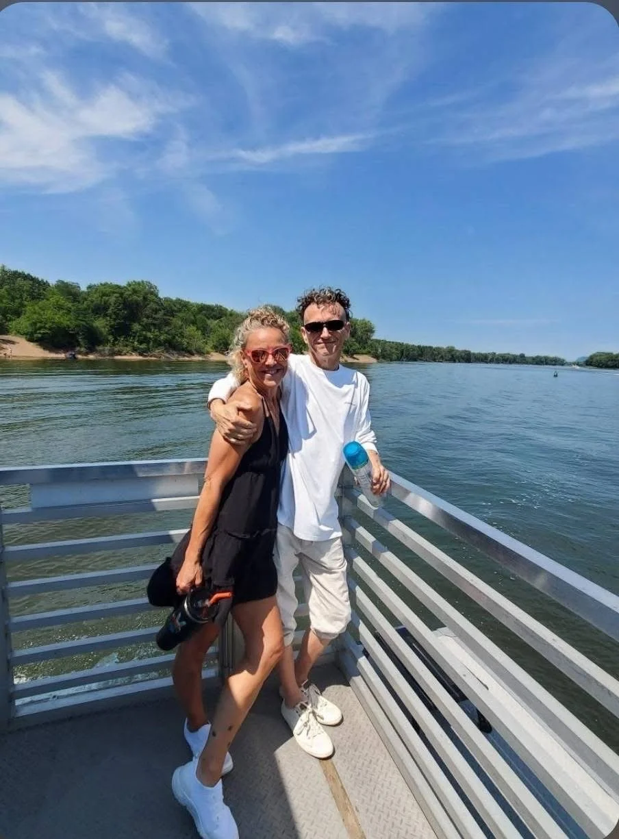 Two people, a woman in a black dress and sunglasses and a man in white clothes and sunglasses, standing close together on a boat with a river and green trees in the background on a sunny day.