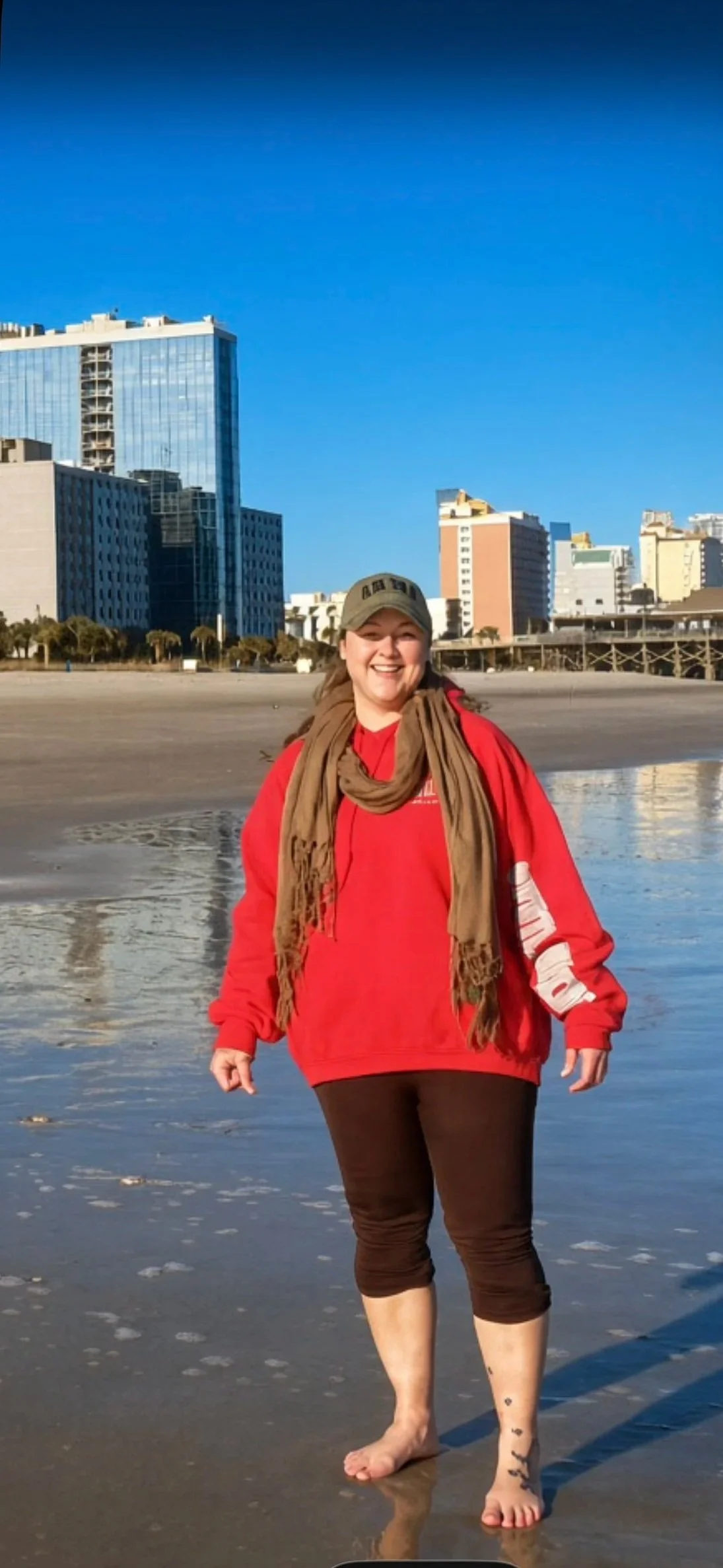 A woman smiling on a beach with skyscrapers in the background. She is barefoot, wearing a red sweatshirt, a brown scarf, and a baseball cap.