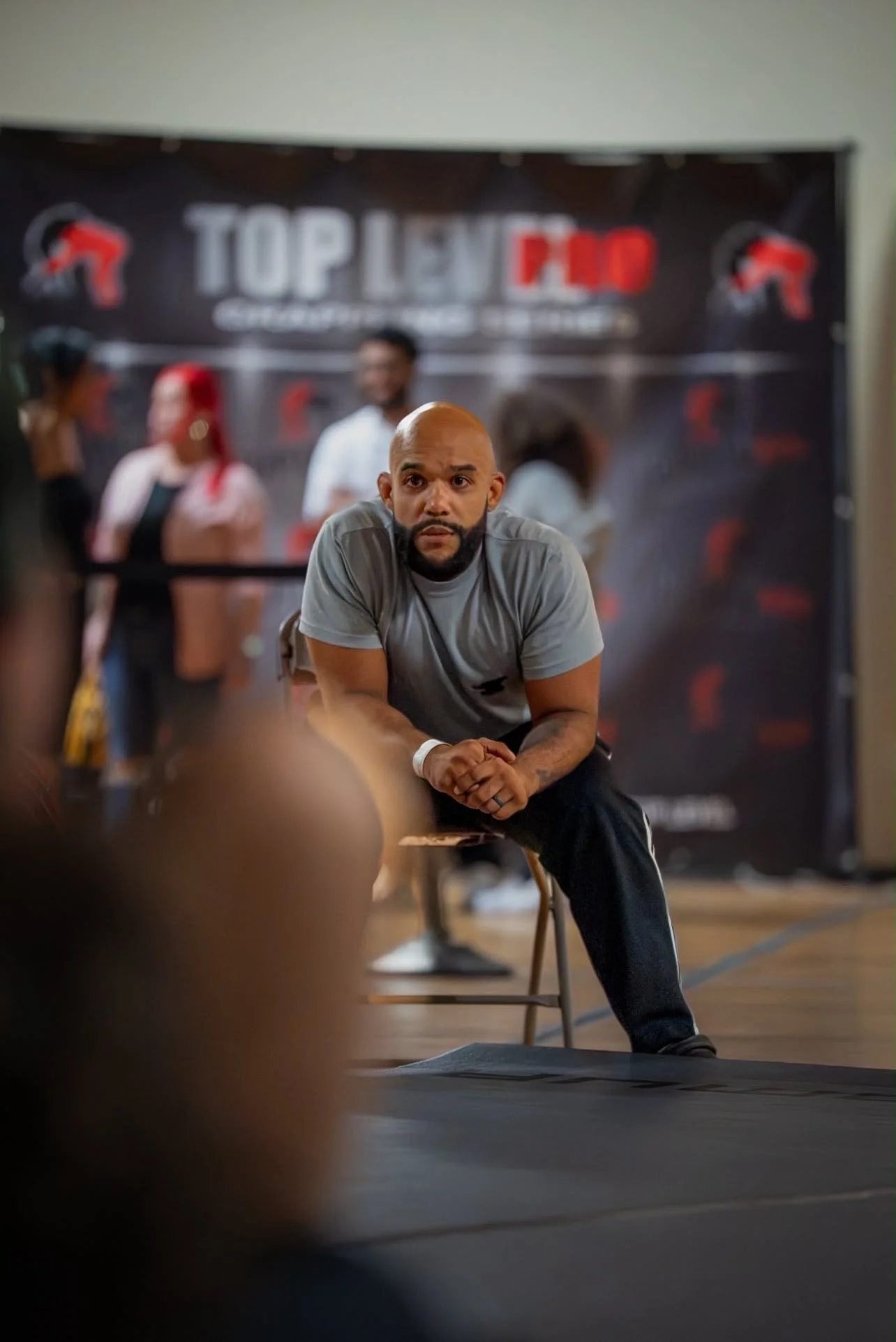 A man with a bald head, beard, and gray t-shirt sitting on a chair in front of a black backdrop with the words "Top Level" and other text, with blurred people in the background.