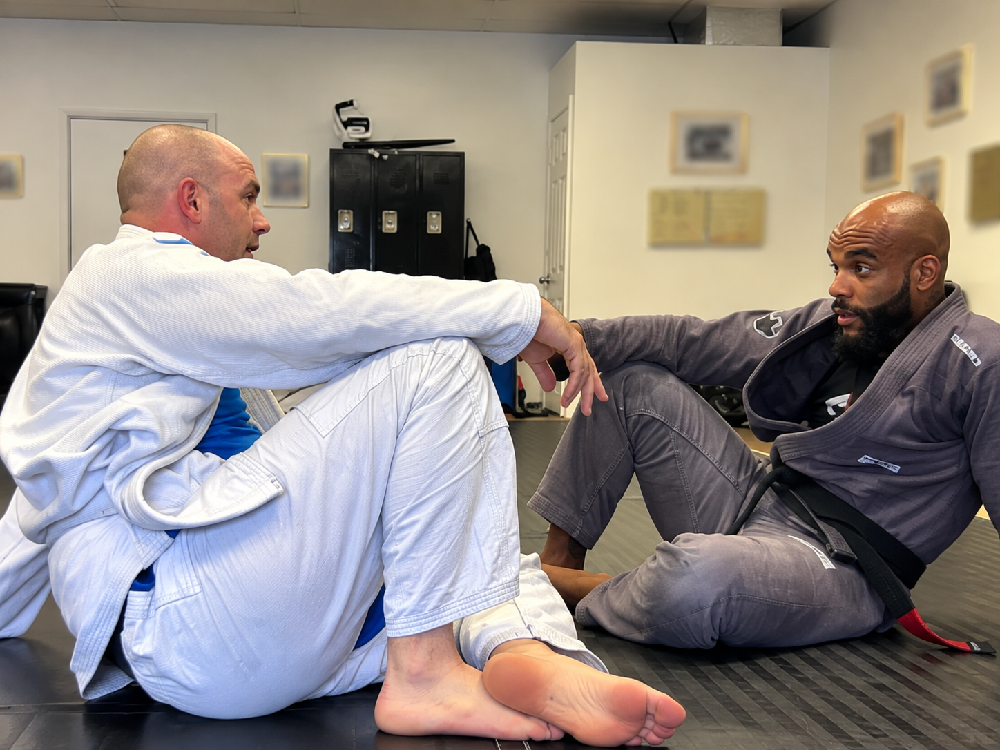 Two men practicing Brazilian Jiu-Jitsu on a black mat in a dojo. One man is in a white gi, sitting with his knees bent, and the other man is in a gray gi with a black belt, sitting across from him. They appear to be engaged in a training or sparring session.