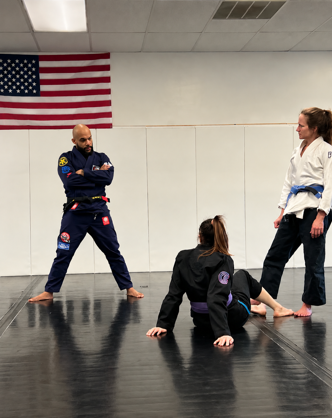 Brazilian Jiu Jitsu black belt instructor coaching two women during a training session, with one student seated on the mat and another standing nearby.