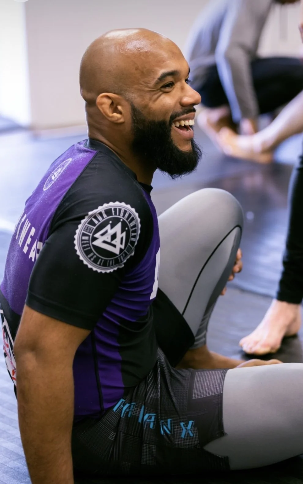 A man with a bald head, full beard, and smiling face, wearing athletic clothing and sitting on the floor. There are other people in the background, and the setting appears to be a gym or training facility.