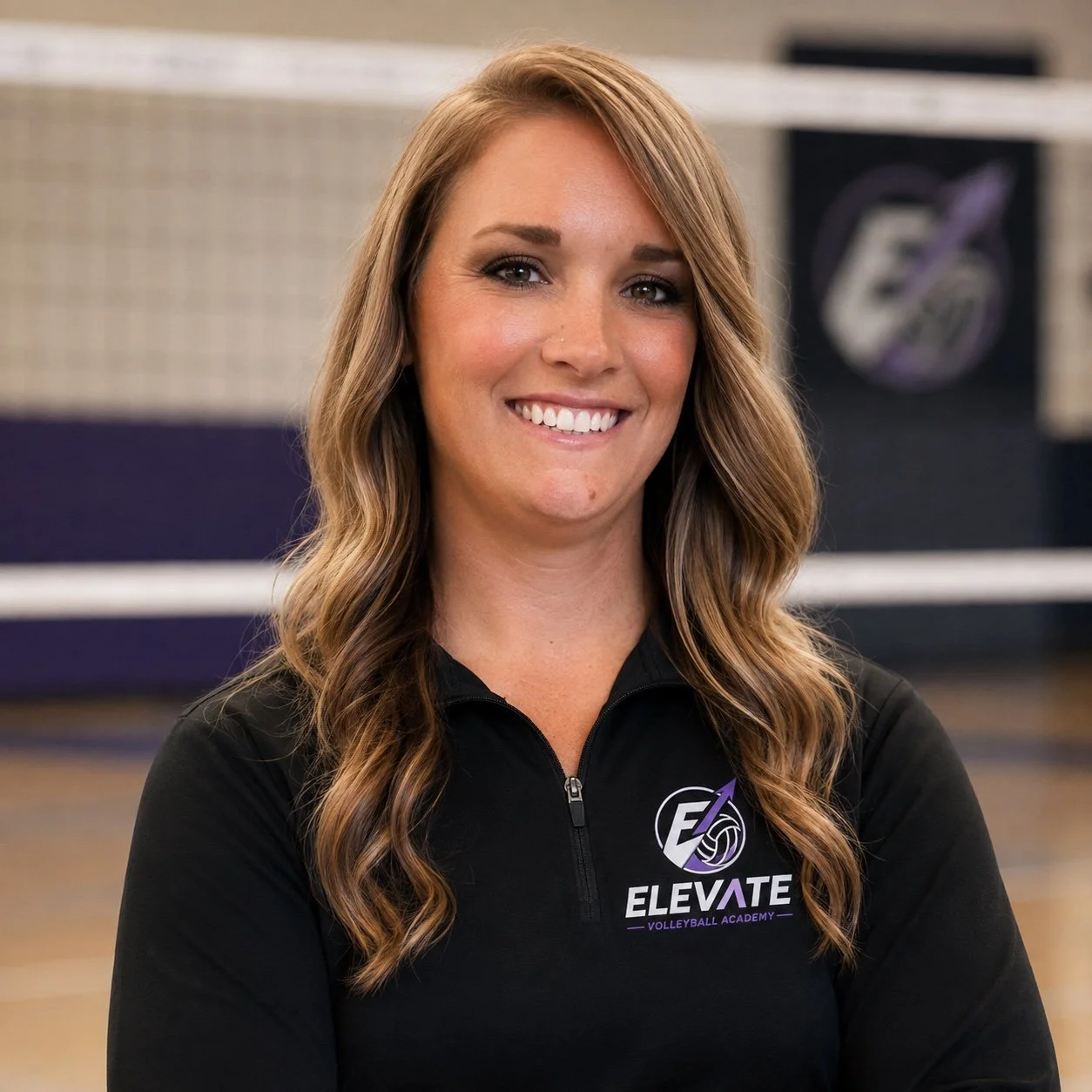 A woman with long wavy brown hair smiling in an indoor volleyball court wearing a black zip-up jacket with the ELIVATE Volleyball Academy logo.