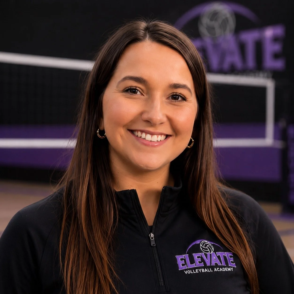 A smiling young woman with long brown hair, wearing a black zip-up jacket with the Elevate Volleyball Academy logo, standing in front of a volleyball net at an indoor volleyball court.