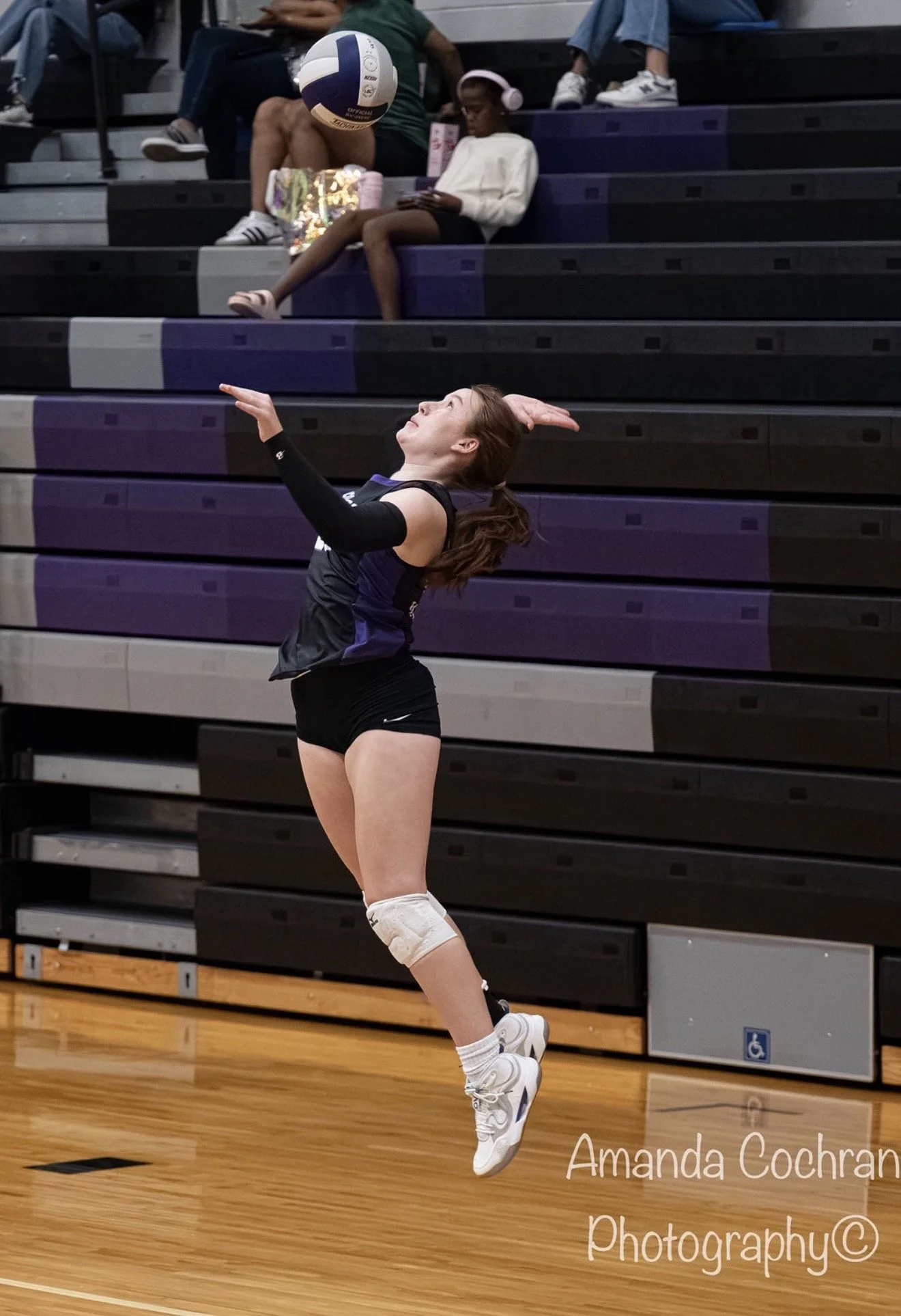 A female volleyball player in mid-air preparing to hit the ball during a game in a gymnasium with black and purple bleachers.