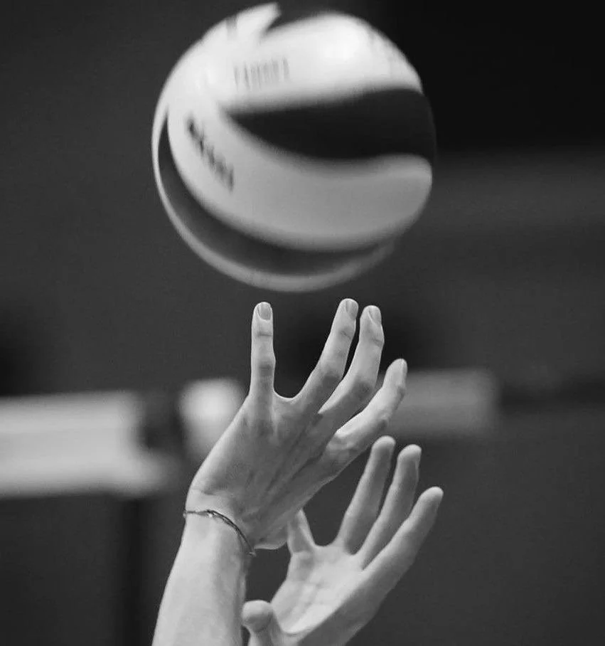 Black and white photo of a volley ball in motion above two hands, with fingers spread apart, reaching out to the ball.