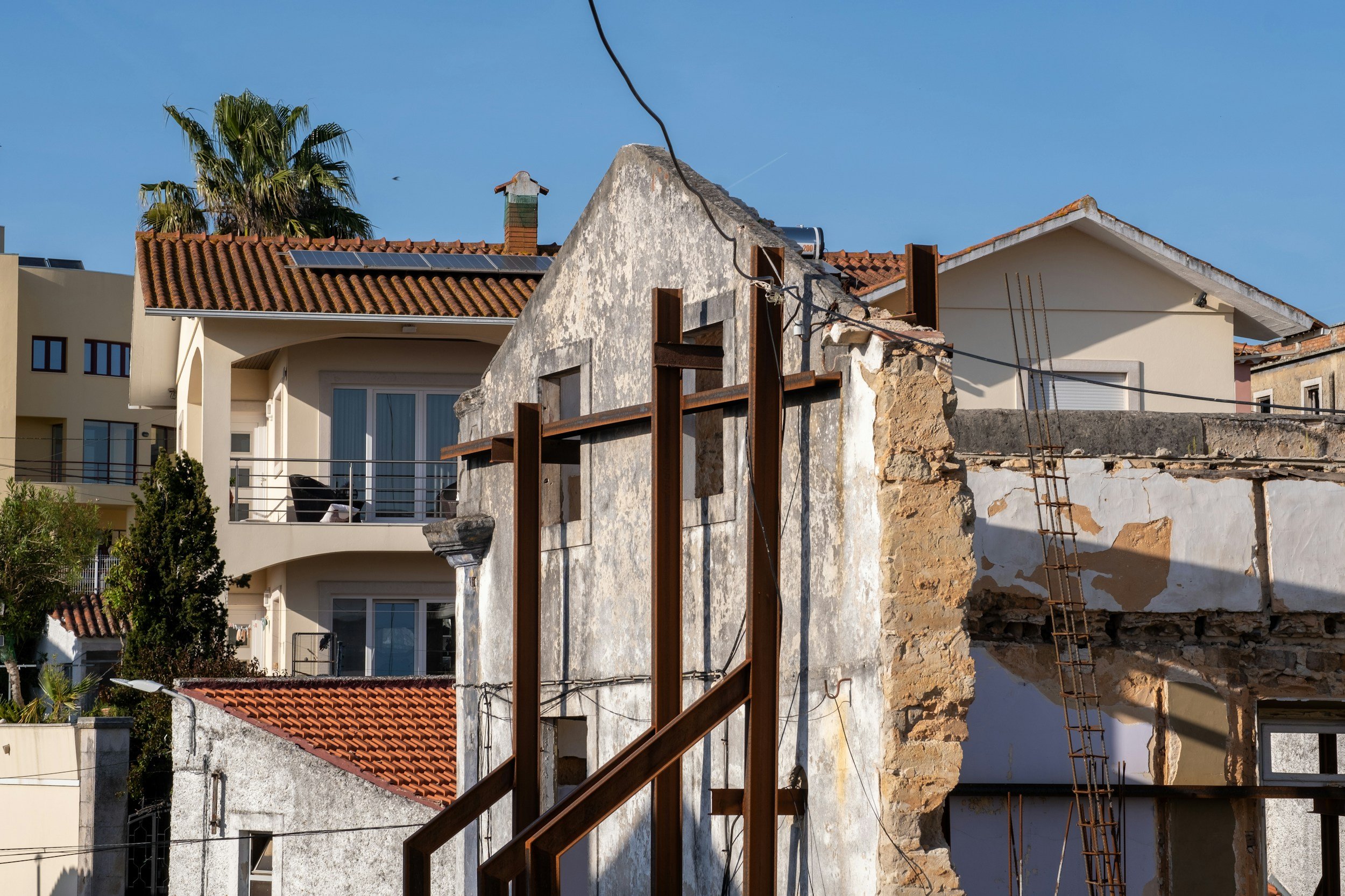 Partially demolished building with exposed brick walls and steel reinforcements, neighboring modern houses, blue sky, and a palm tree.