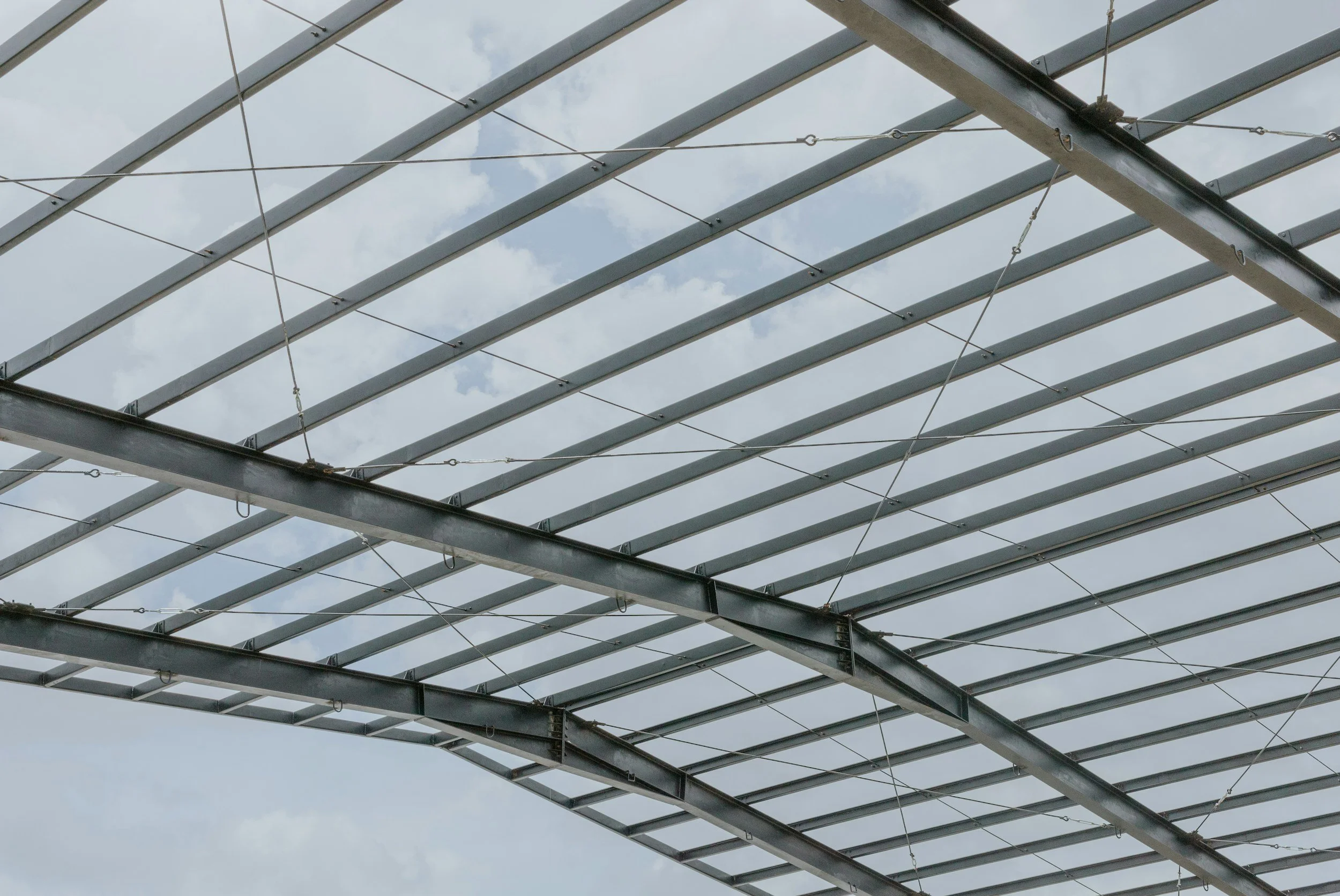 Metal framework of a roof under construction with steel beams and support wires against a cloudy sky.