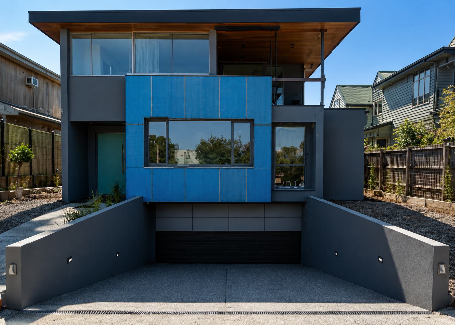 Modern two-story house with a garage below, featuring blue and gray exterior walls, large windows, and a wooden ceiling on the upper balcony, surrounded by neighboring houses and a fence.