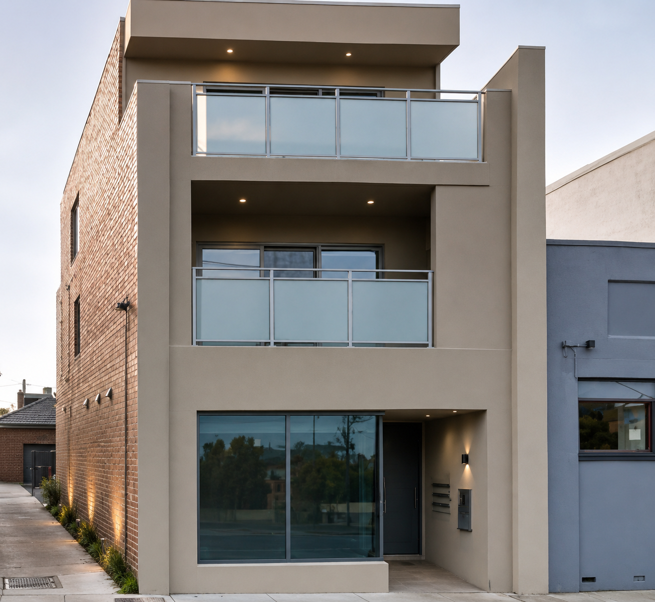 Modern multi-story apartment building with beige and brick exterior, glass balcony railings, and large windows, with small plants along the sidewalk.