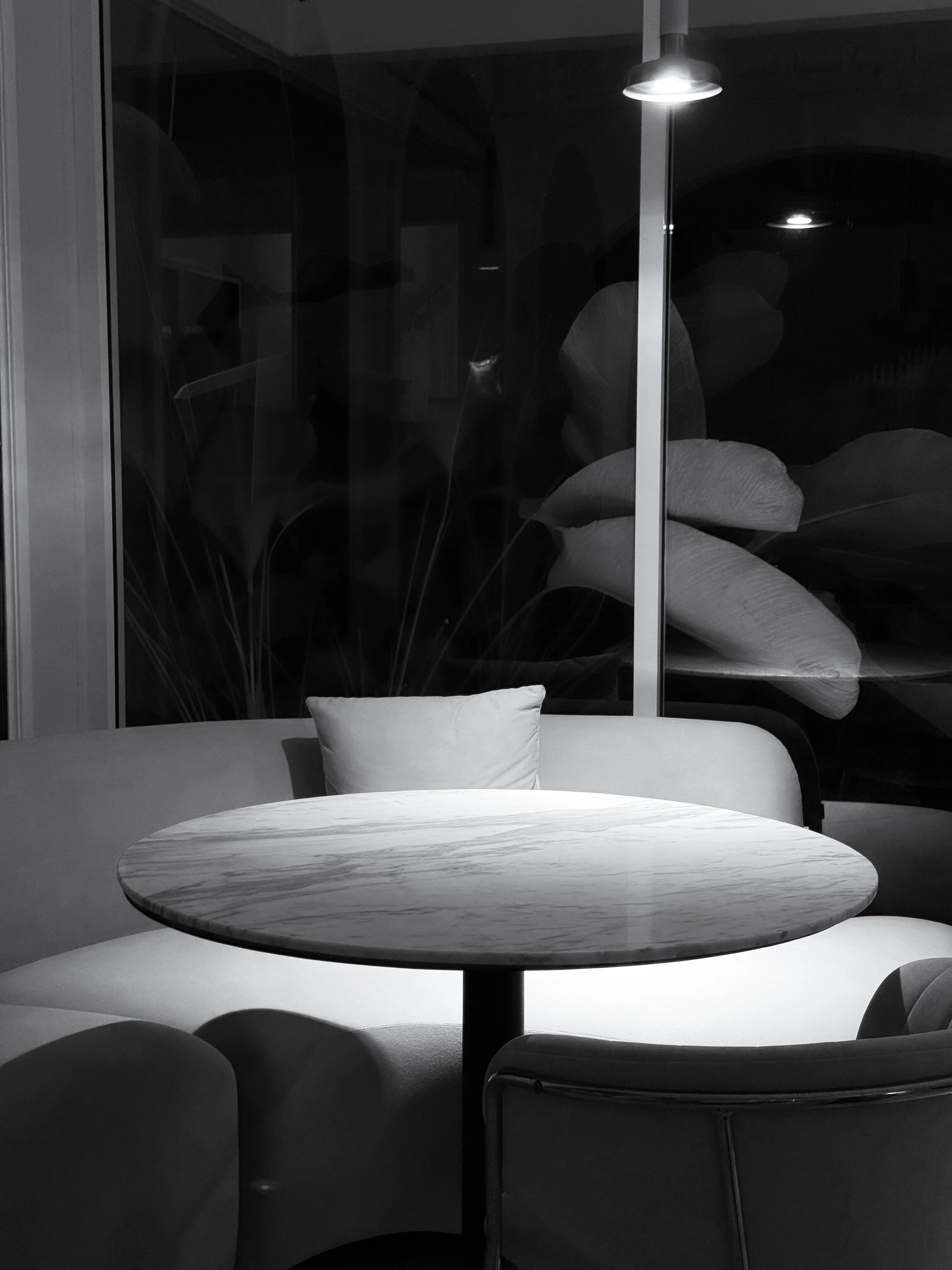 A dining area at night with a round wooden table, white cushioned chairs, and a large window with a glass door. Outside the window, plants and a dark sky are visible. A ceiling light illuminates the scene.