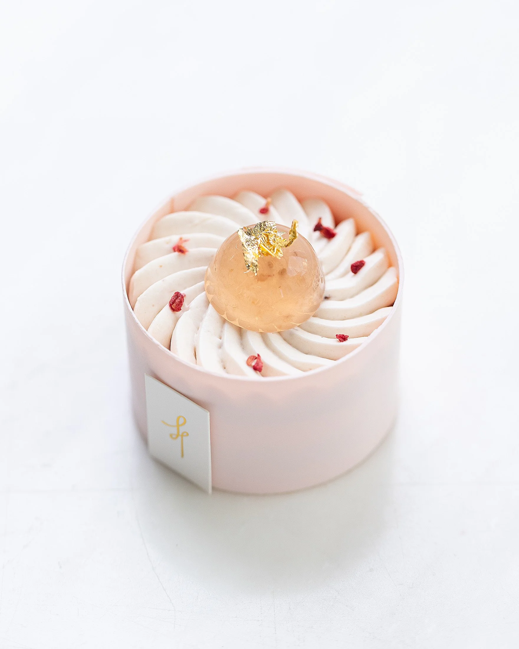 A round, pink box of cupcakes topped with swirled white frosting, small red toppings, and a golden decorative topper, with a white label featuring the initials 'LF'.