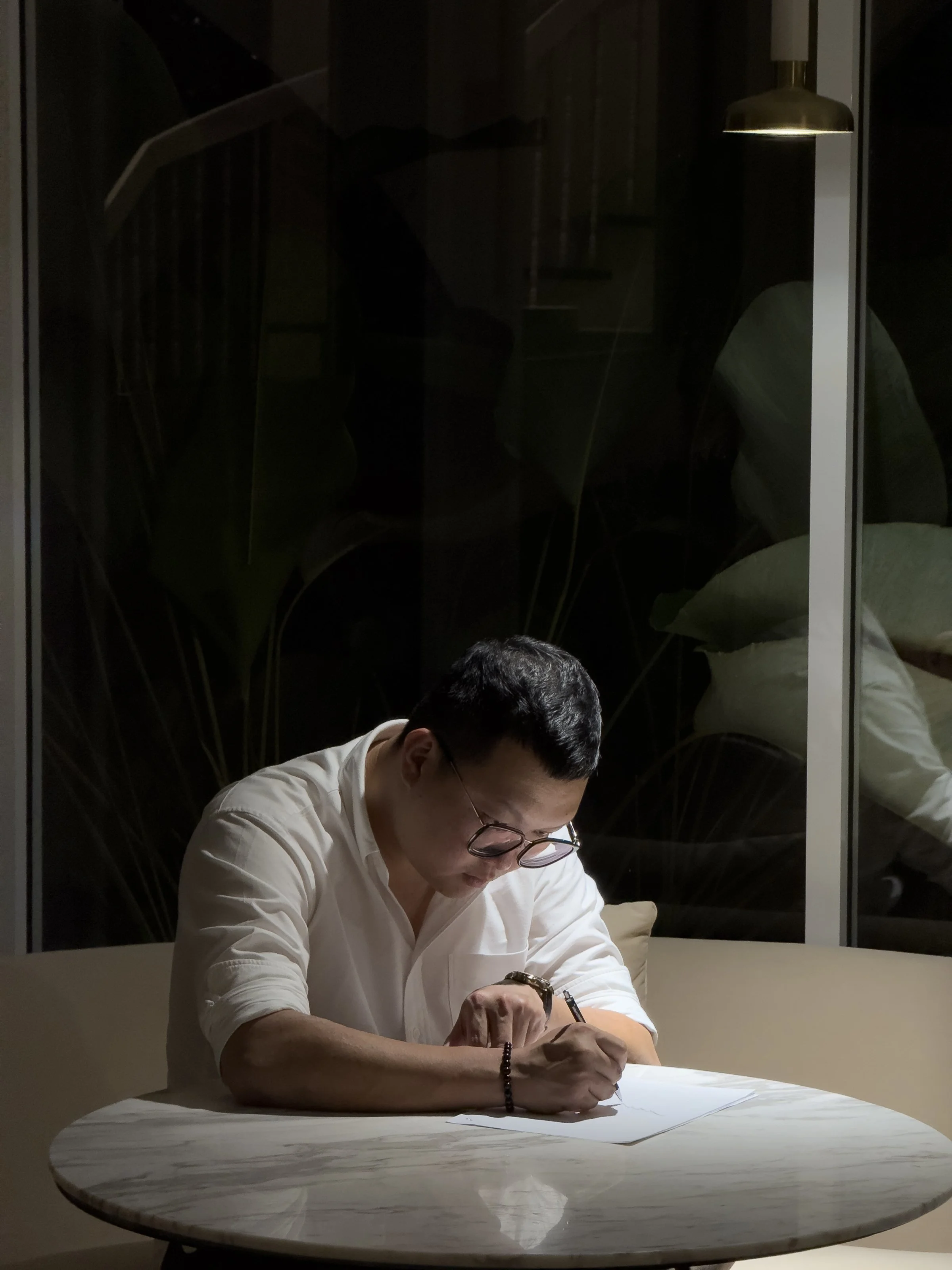 A man with glasses and a white shirt writing on paper at a round marble table, with reflections of him and a lamp in the glass window behind him.