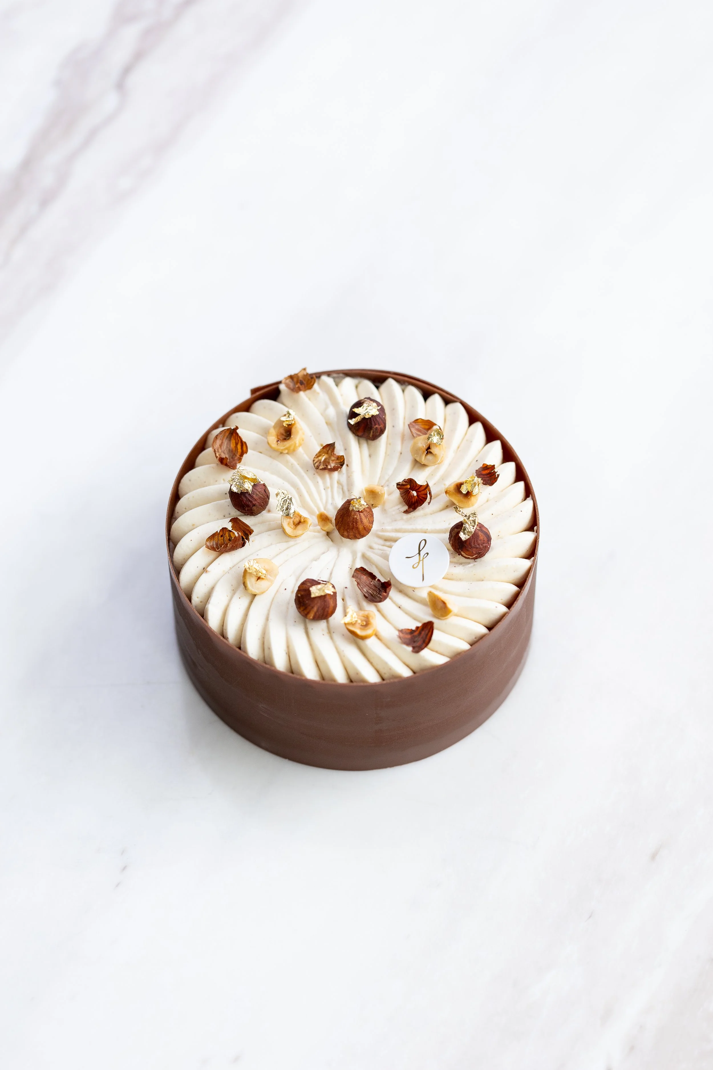 Round cake with white frosting, topped with hazelnuts and gold leaf, in a brown oval box on a white marble surface.