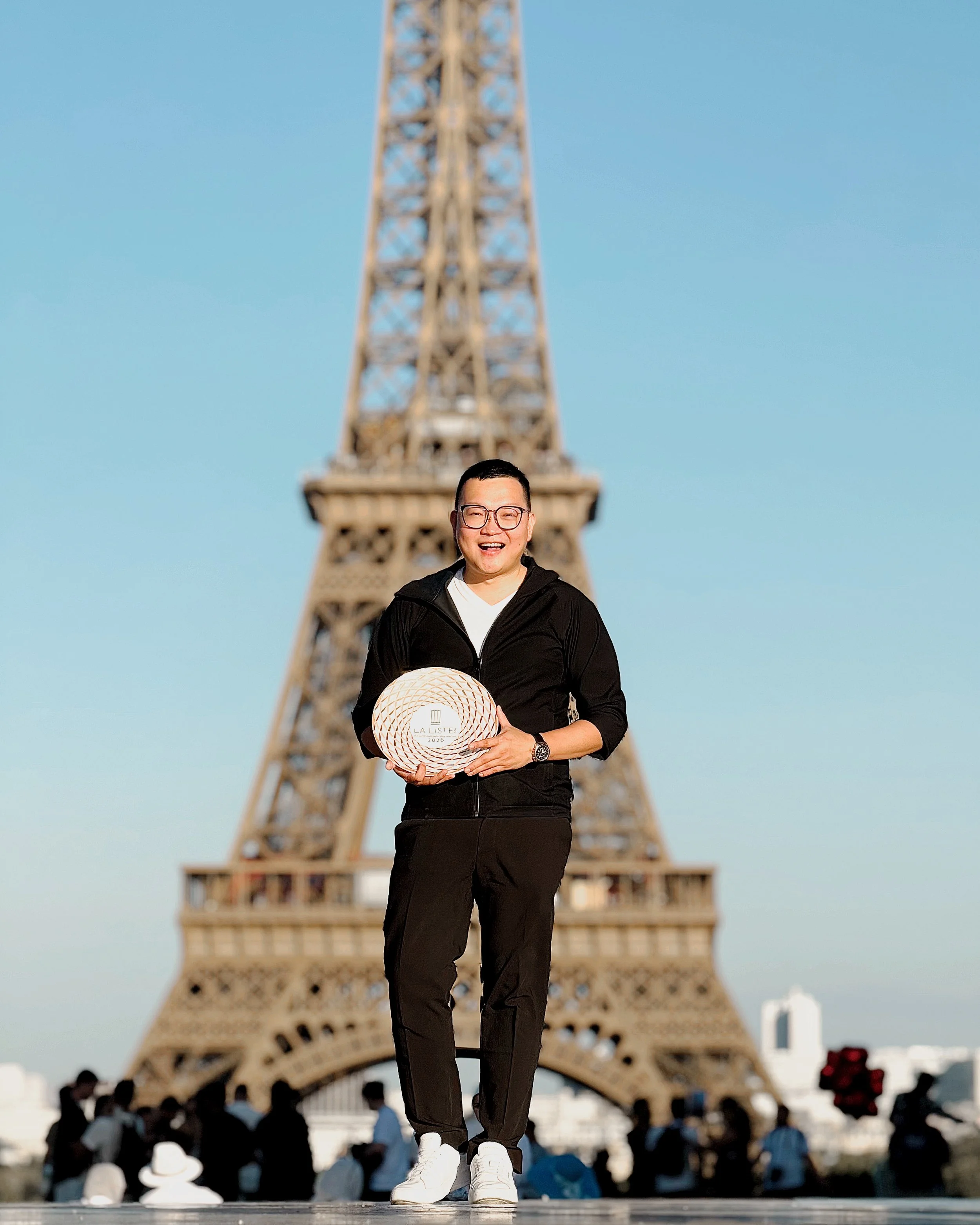 A man smiling wearing glasses and holding a white circular fan with words in French, standing in front of the Eiffel Tower during daytime.