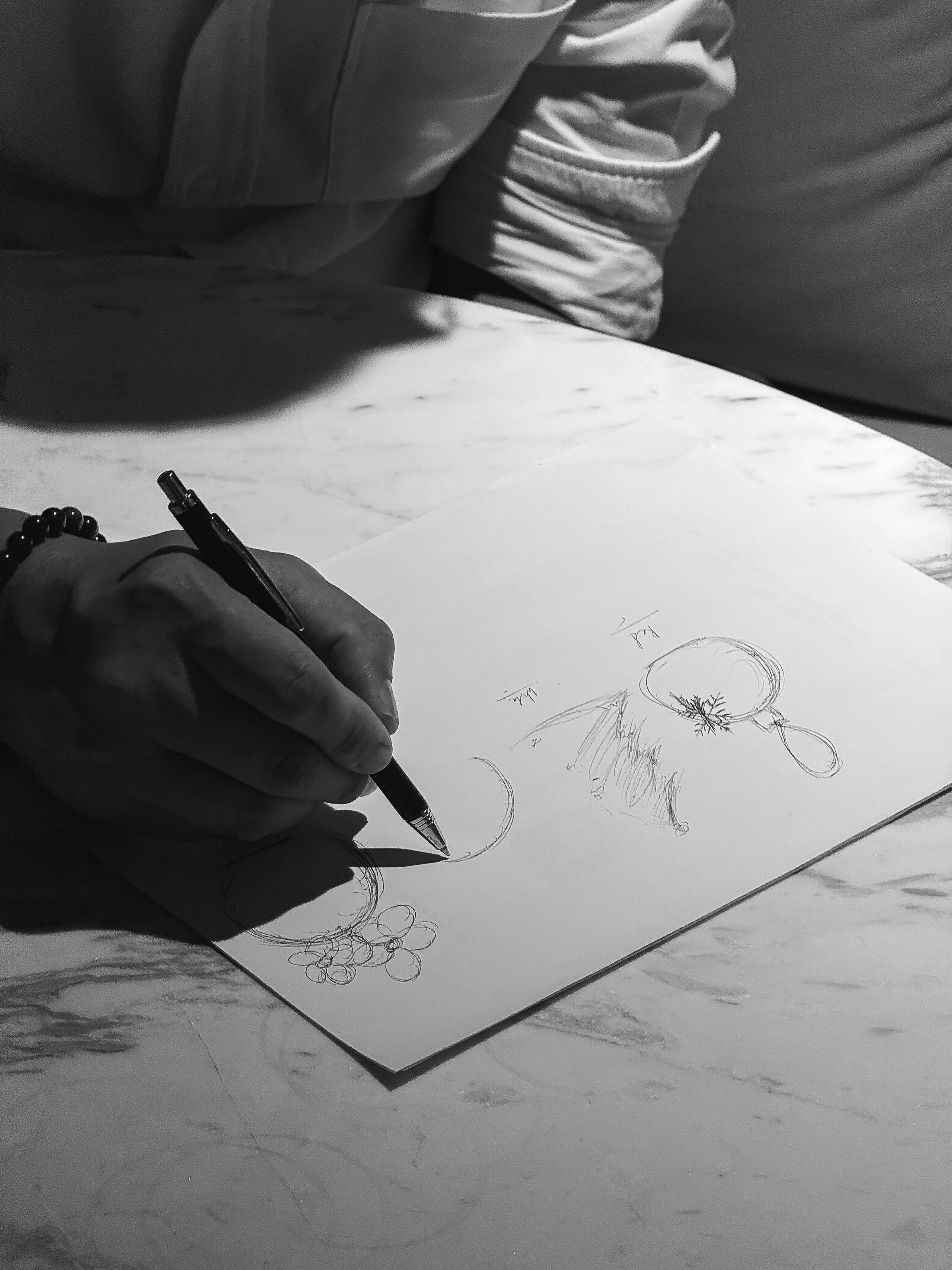 A person sketching a scene of animals and trees on a sheet of paper, with another person sitting nearby. The scene is black and white.