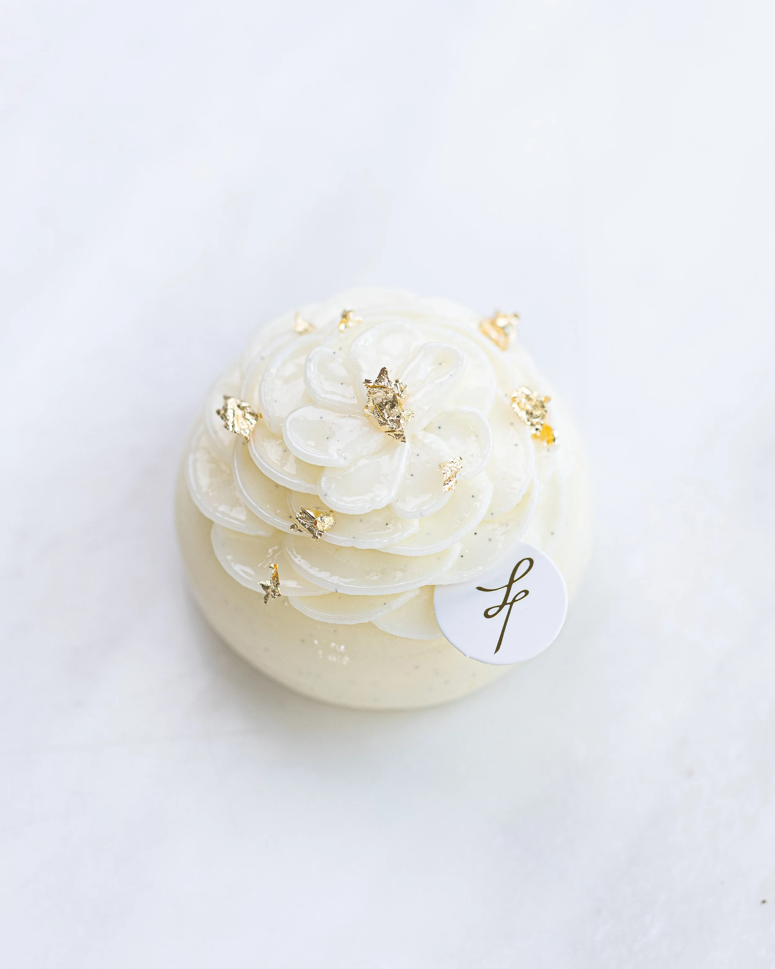 A white dessert topped with white chocolate curls, edible gold leaf, and a small white tag with a gold cursive letter 'L' on it, set against a white background.
