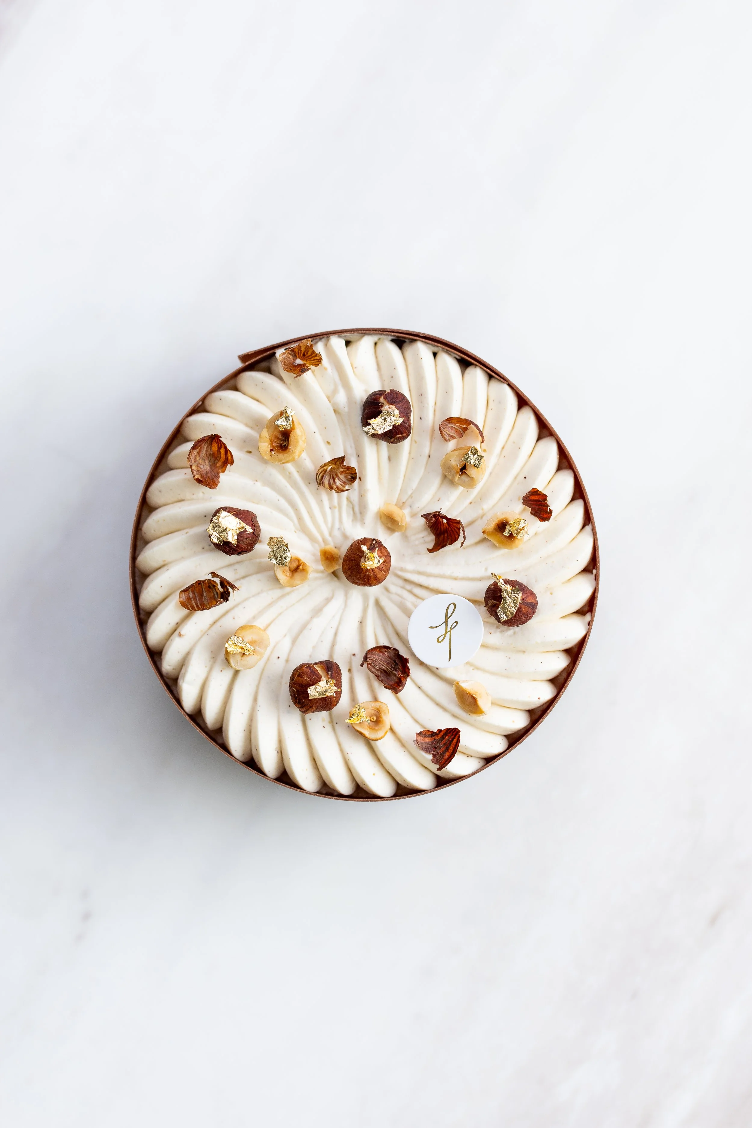 Round dessert topped with piped white frosting, hazelnuts, and gold leaf accents, presented against a white background.