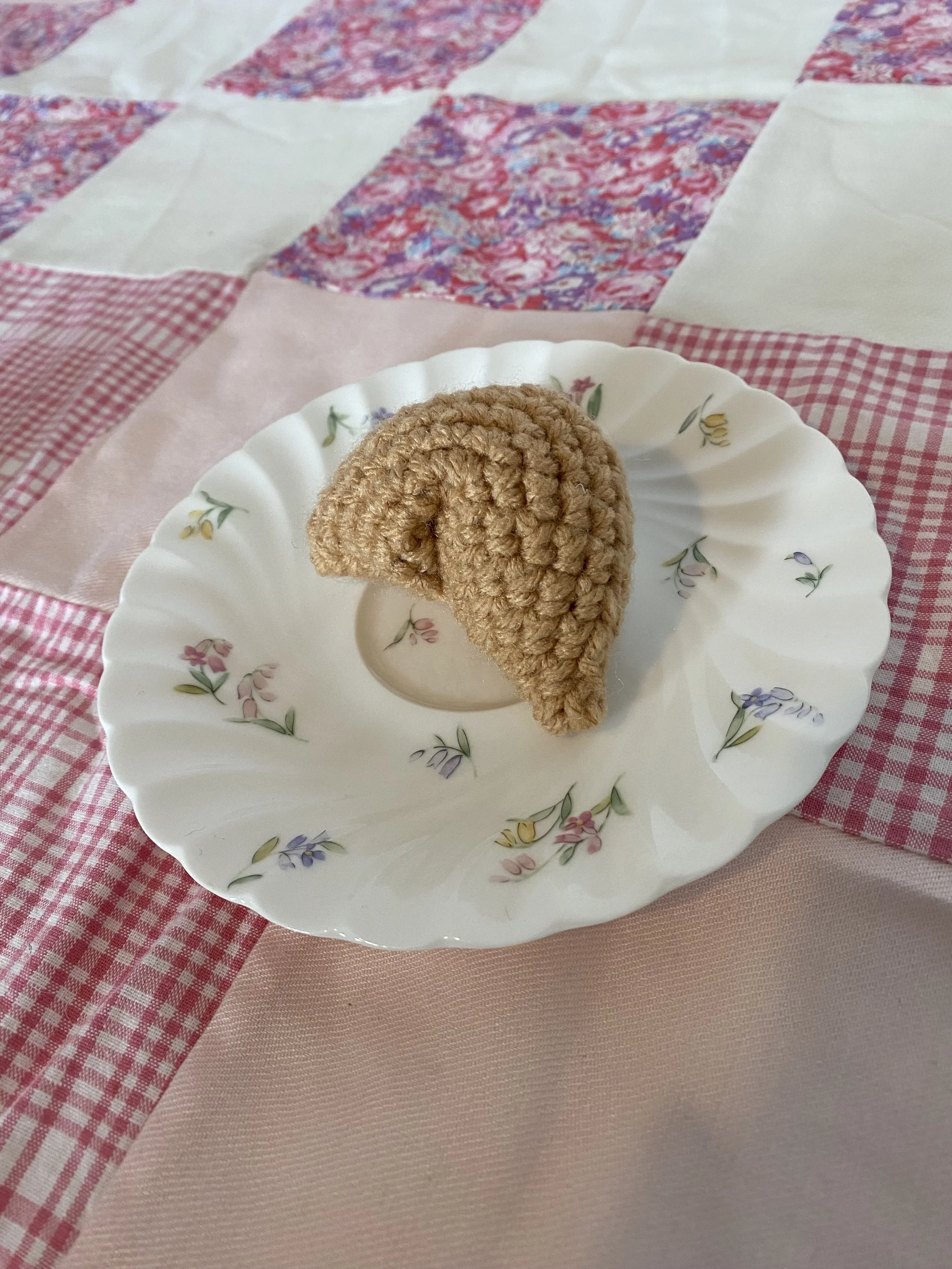 A crocheted fortune cookie on a floral pattern plate, set on a pink and red checkered tablecloth with a pink solid tablecloth underneath.