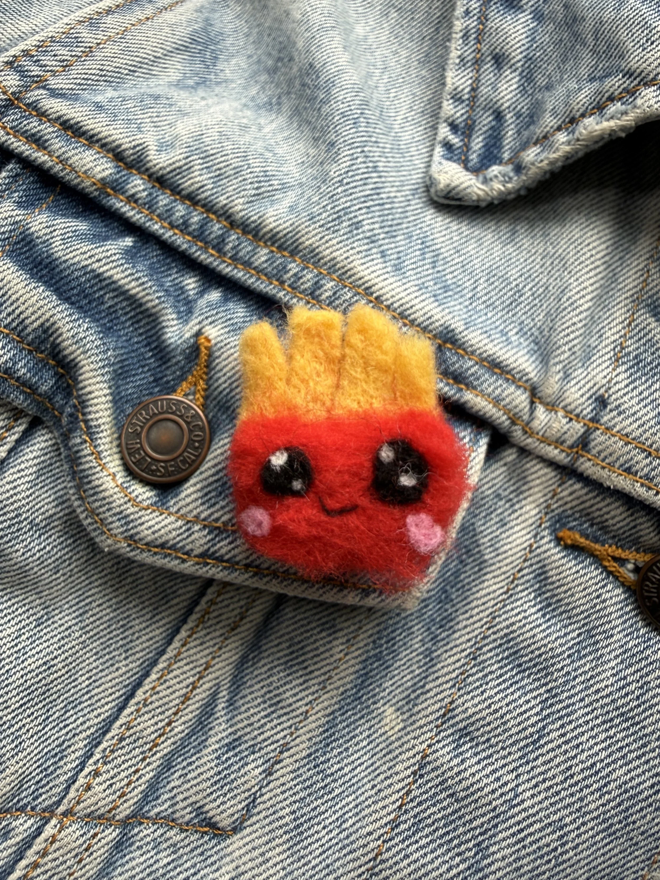 Close-up of a pair of blue denim jeans with a felted french fries pin on the pocket.