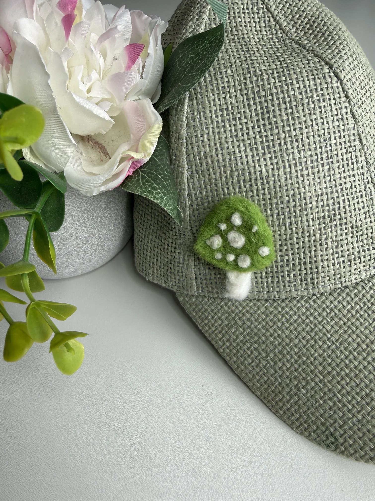 Artificial white and pink flower with green leaves, a green felted mushroom, and a woven basket on a white surface.