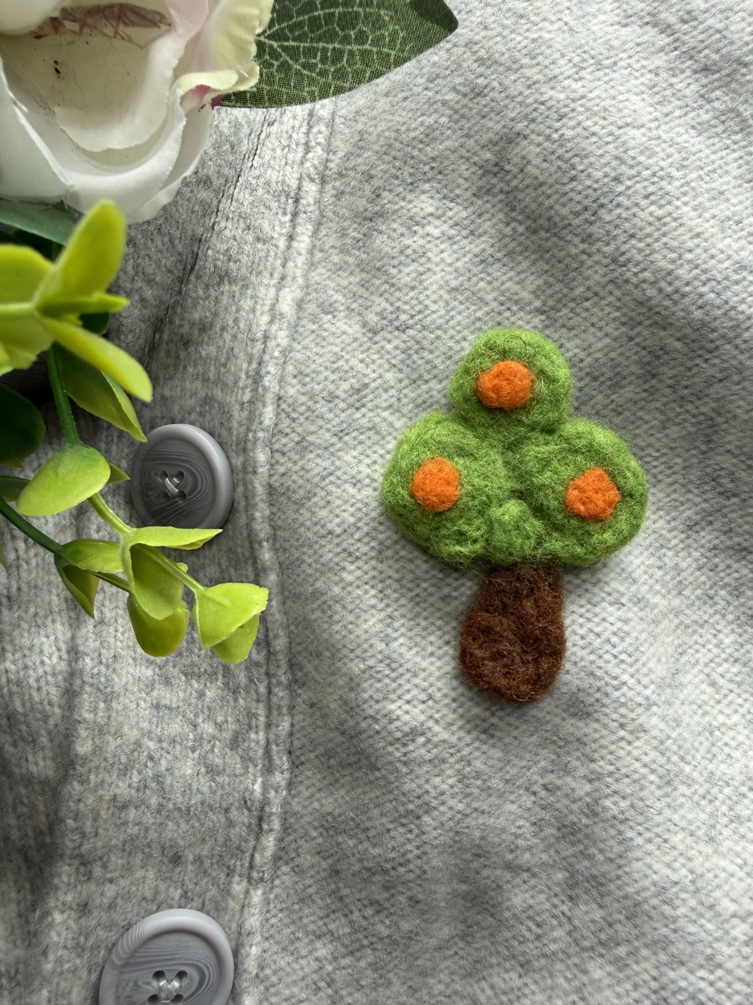 Close-up of an Animal Crossing orange tree pin, a white flower in a small white pot, a green plant, and a grey button.