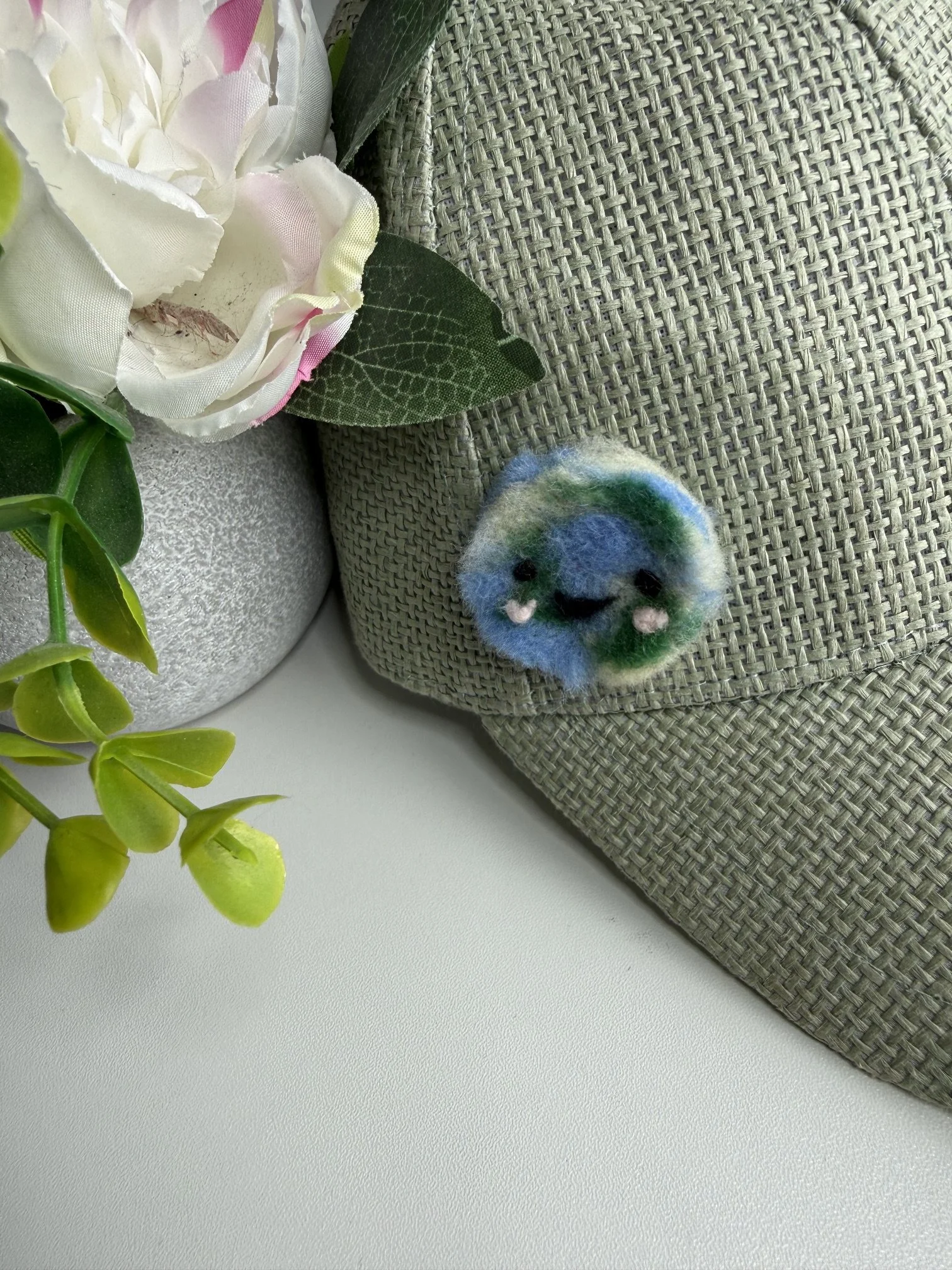 A decorative artificial flower arrangement with white and pink fabric flowers, green fabric leaves, set on a gray textured surface beside a silver vase, partially covering a woven gray placemat with a small felted earth pin.