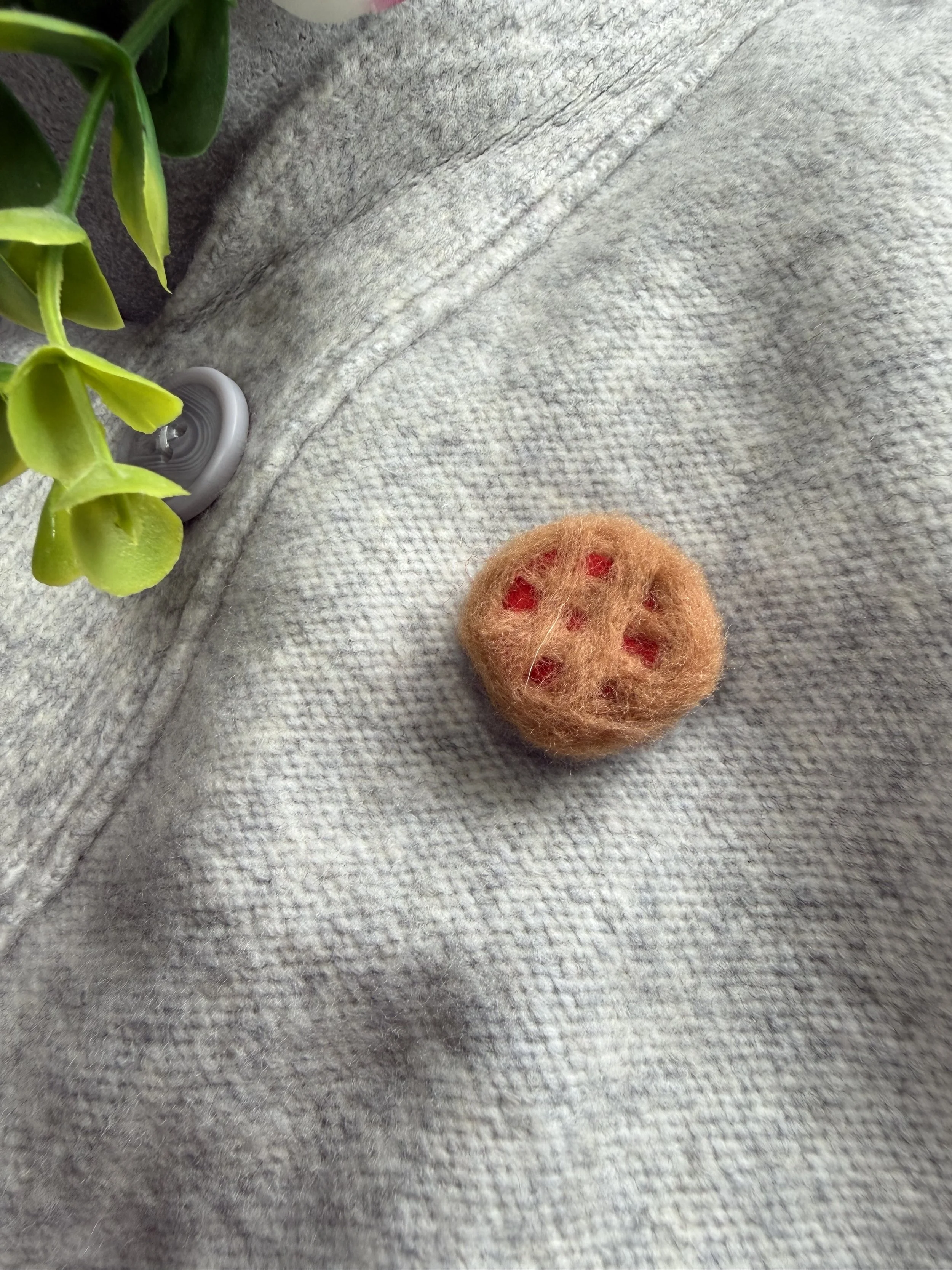 Close-up of a gray fabric surface with a small orange felted pie, green leaves partially visible, and a gray button.