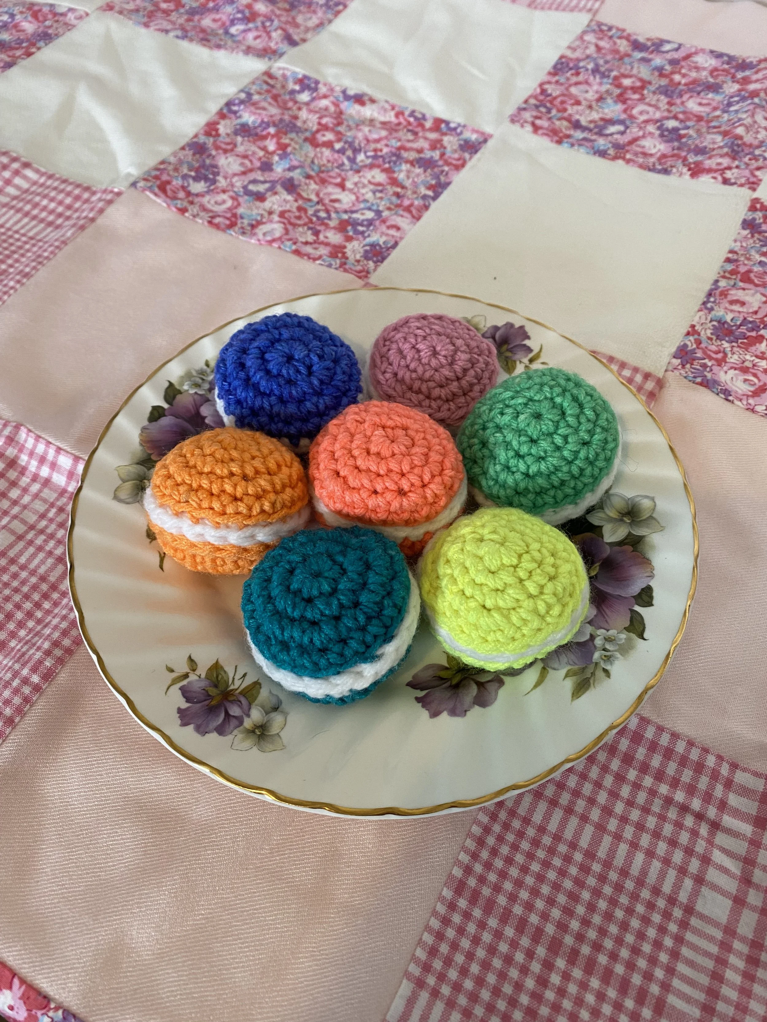 A plate of colorful crochet macarons on a floral and checkered tablecloth.