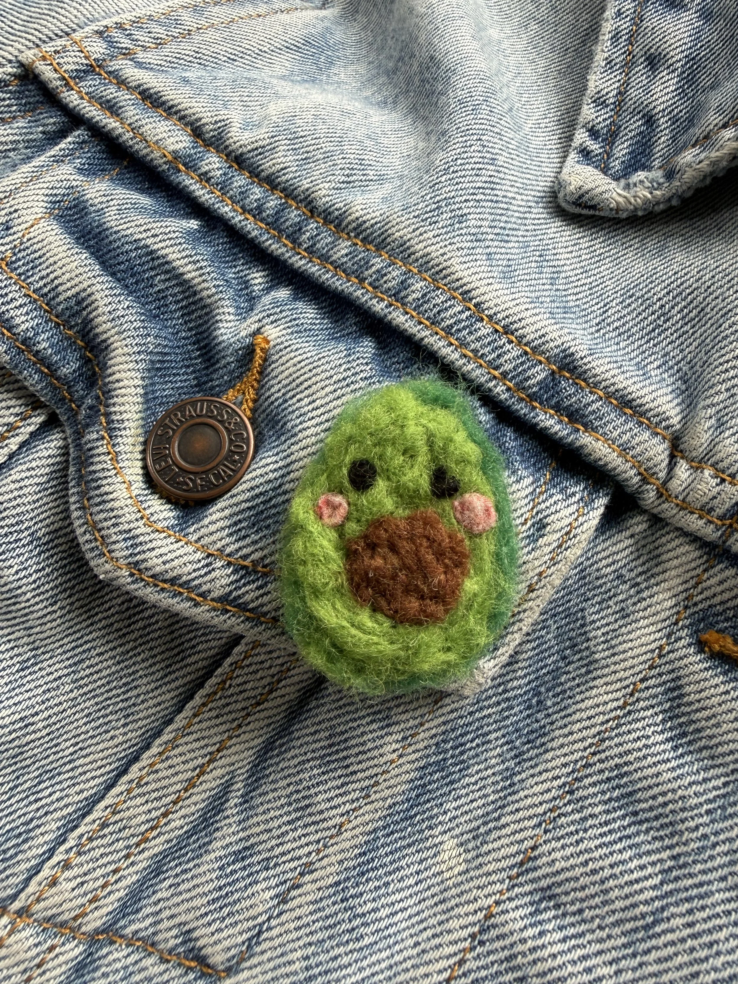 Close-up of a blue denim pocket with orange stitching, featuring a felted green avocado-shaped patch with a brown pit and pink cheeks.