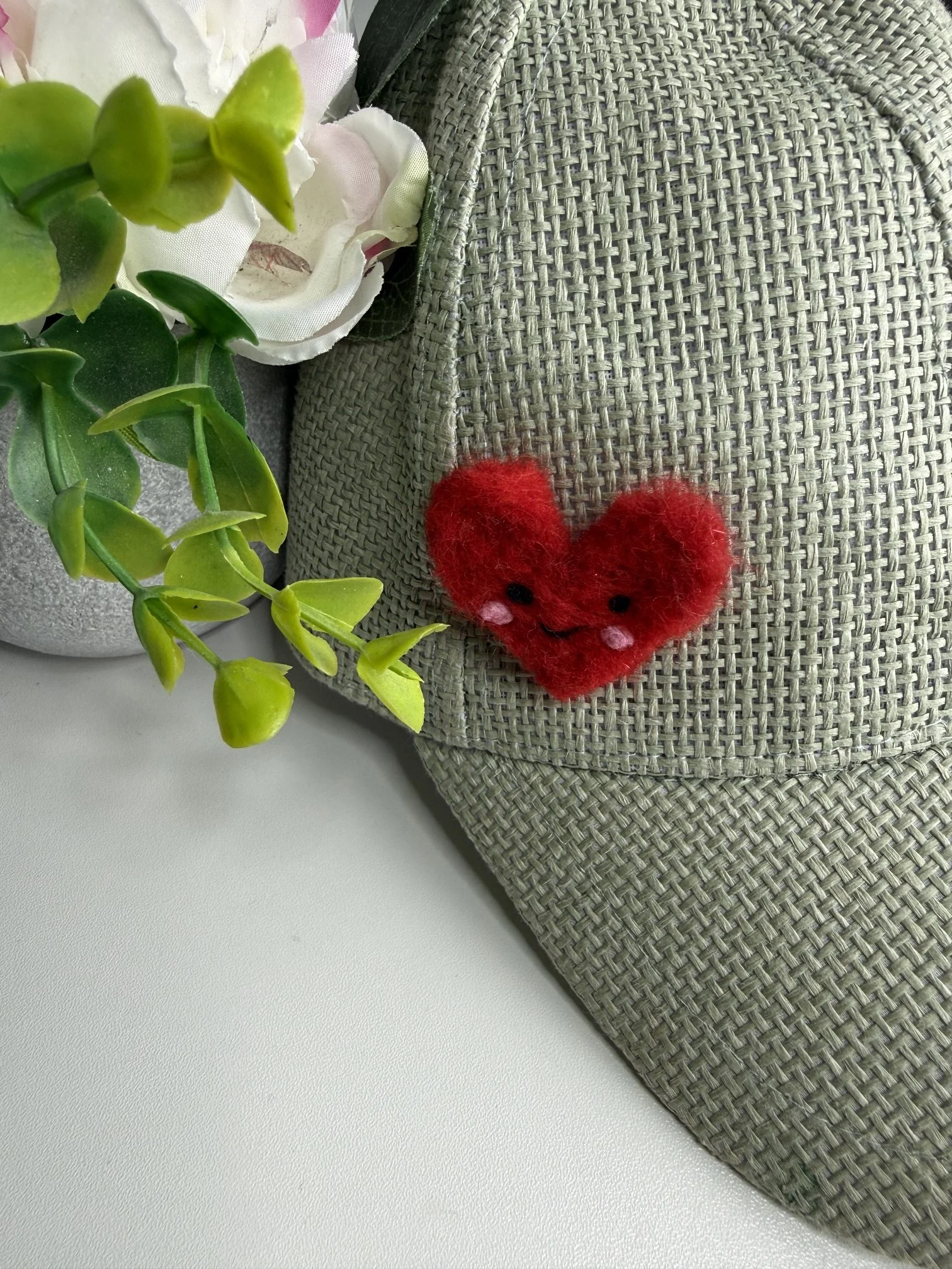 A decorative arrangement featuring a green leafy plant, faux white flowers, and a small felted red fuzzy heart with black eyes, placed on a woven gray placemat on a white surface.
