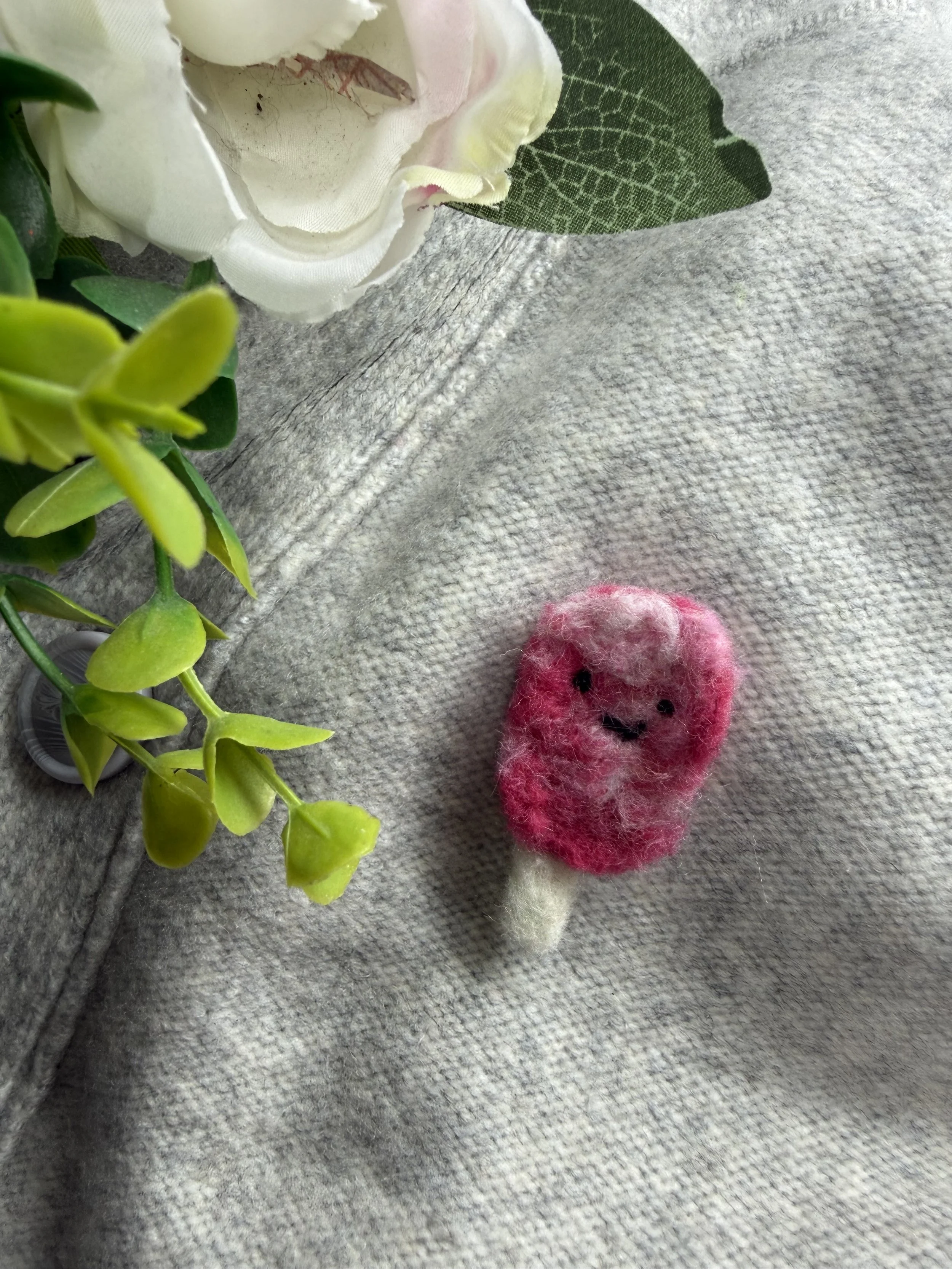 Close-up of a white artificial flower with a green leaf, some greenery, and a small pink felted popsicle.