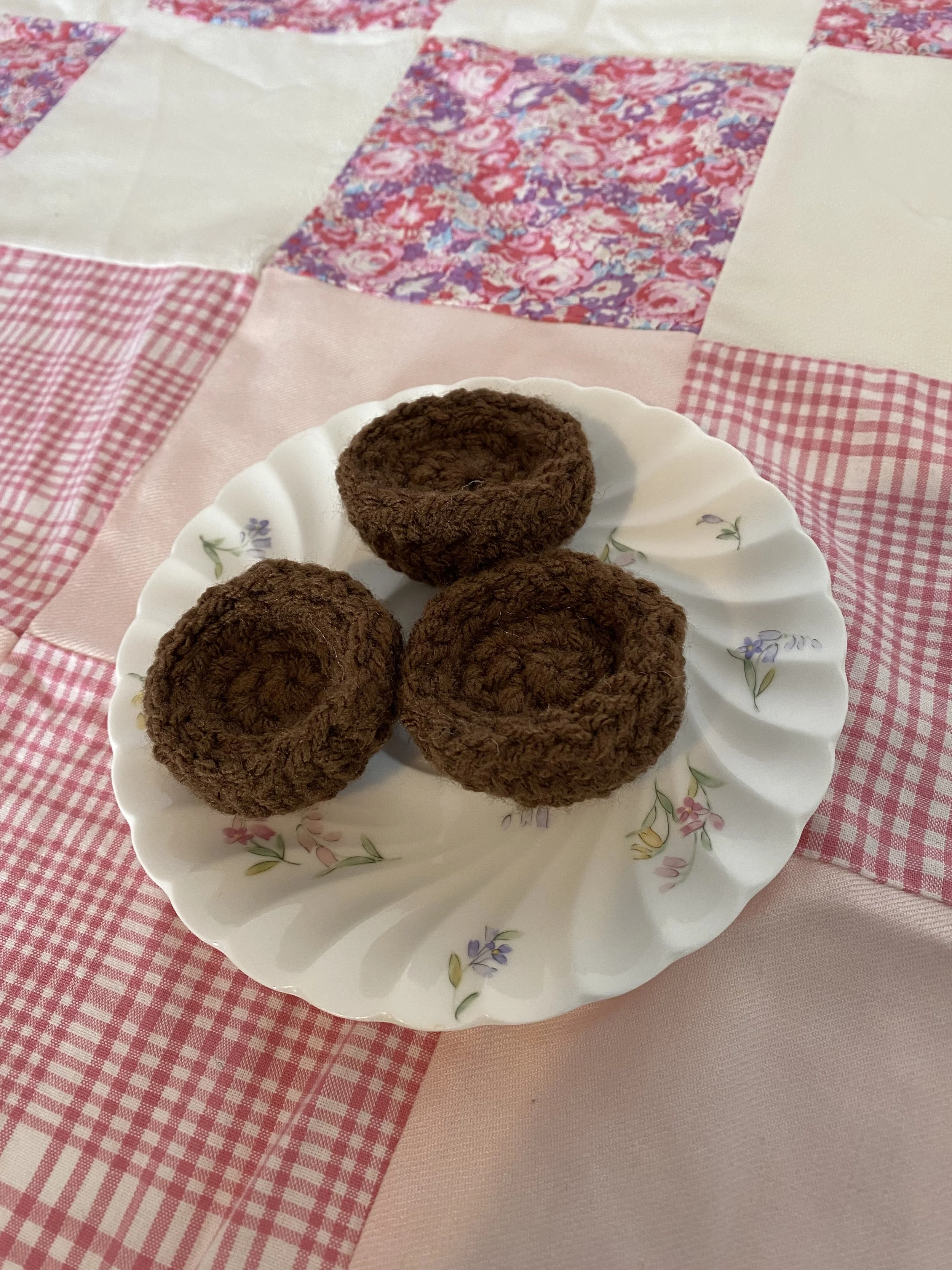 A white ceramic plate with a floral pattern containing crocheted two bite brownies, on a pink checkered and plain tablecloth.
