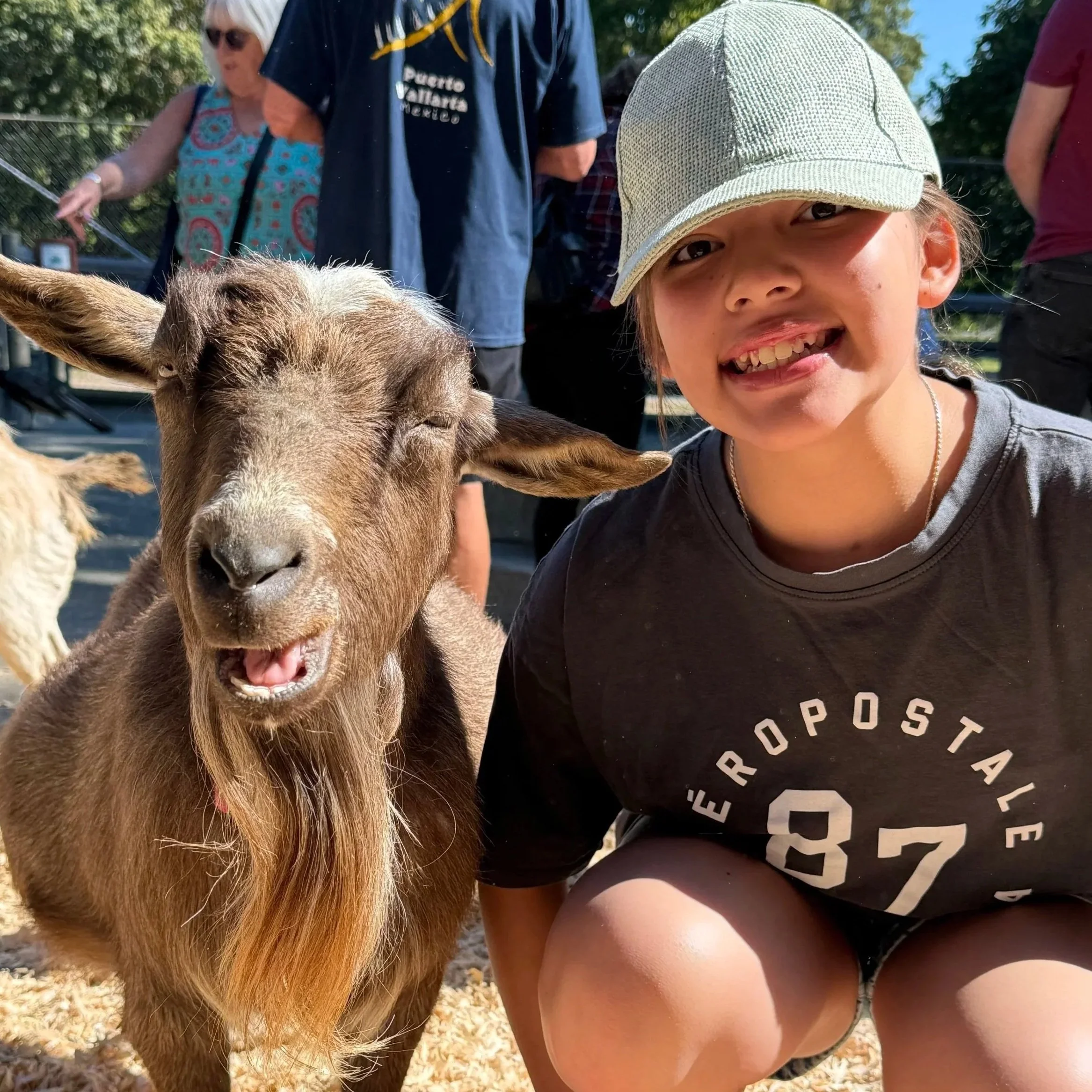 A young girl smiling, wearing a light-colored baseball cap and a gray t-shirt, with a brown and white goat standing next to her. In the background, there are other people outdoors and trees.